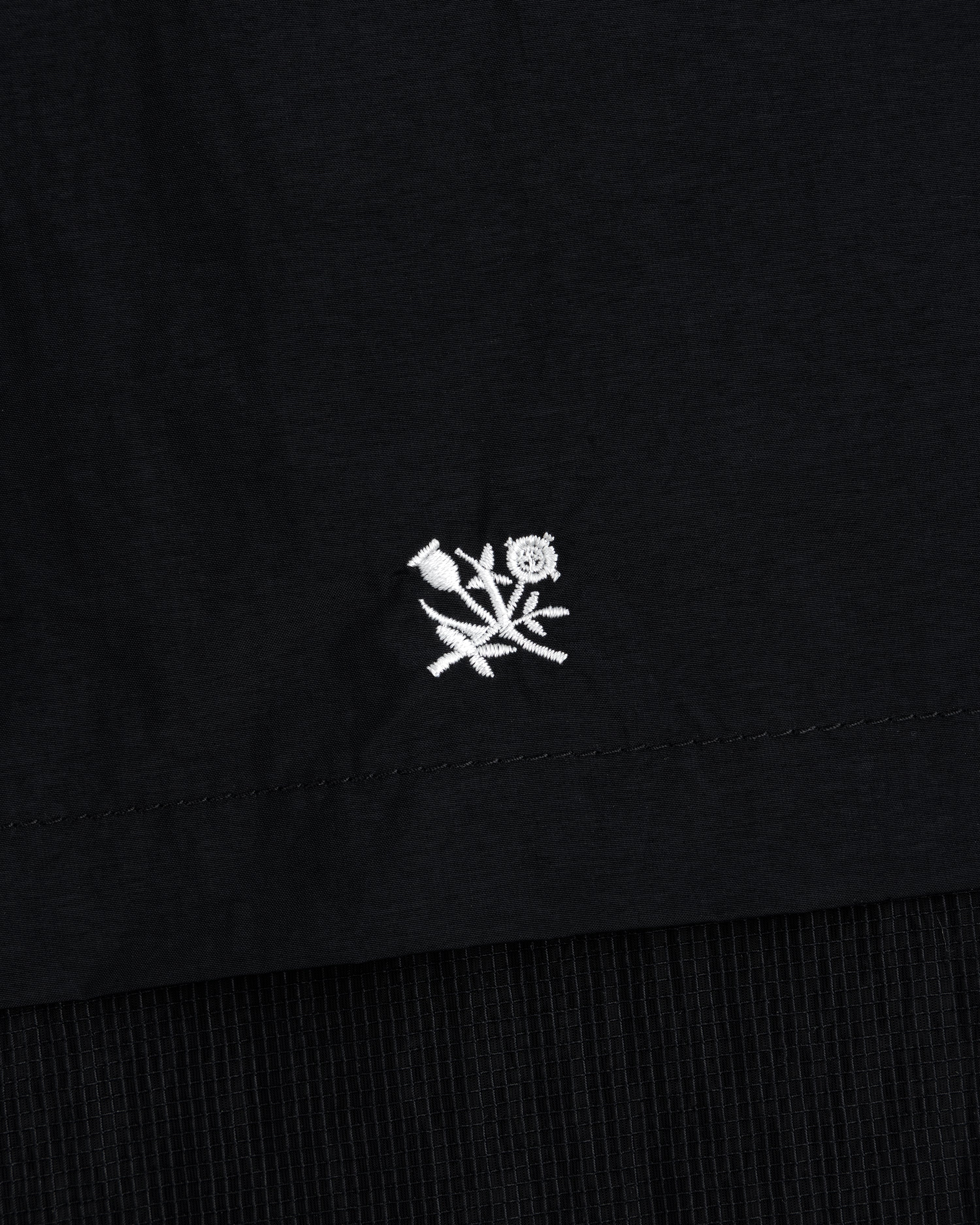 Small white embroidered rose with leaves on black Nylon Track Jacket, near the hem.