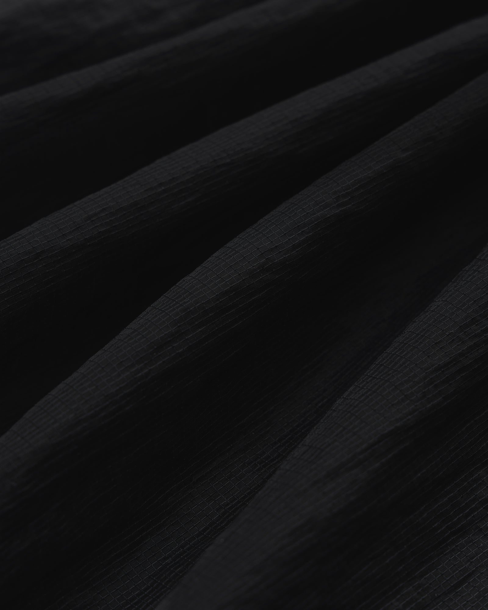 Close-up of black Nylon Track Jacket fabric with a textured, slightly ridged surface and soft, diagonal folds.