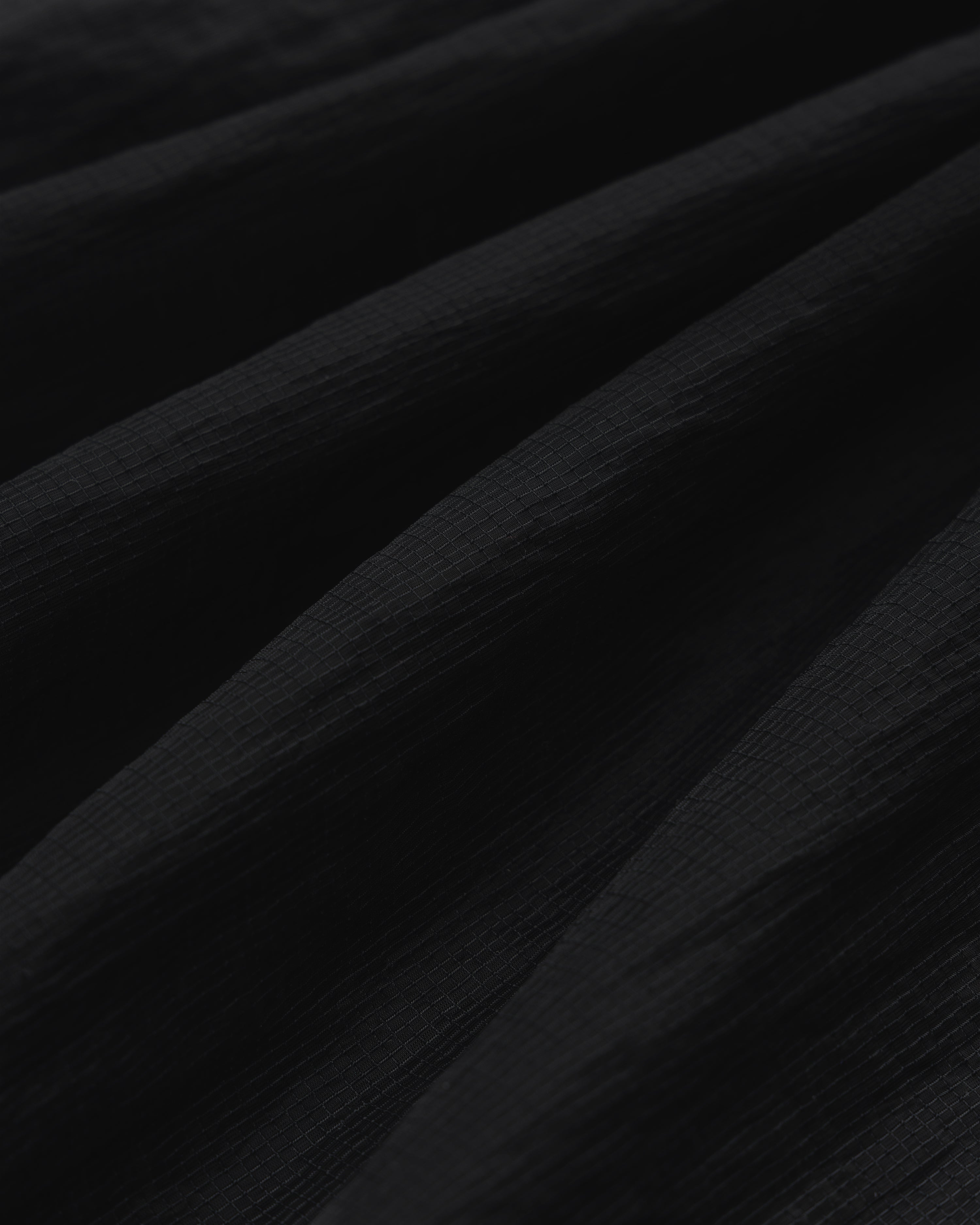 Close-up of black Nylon Track Jacket fabric with a textured, slightly ridged surface and soft, diagonal folds.