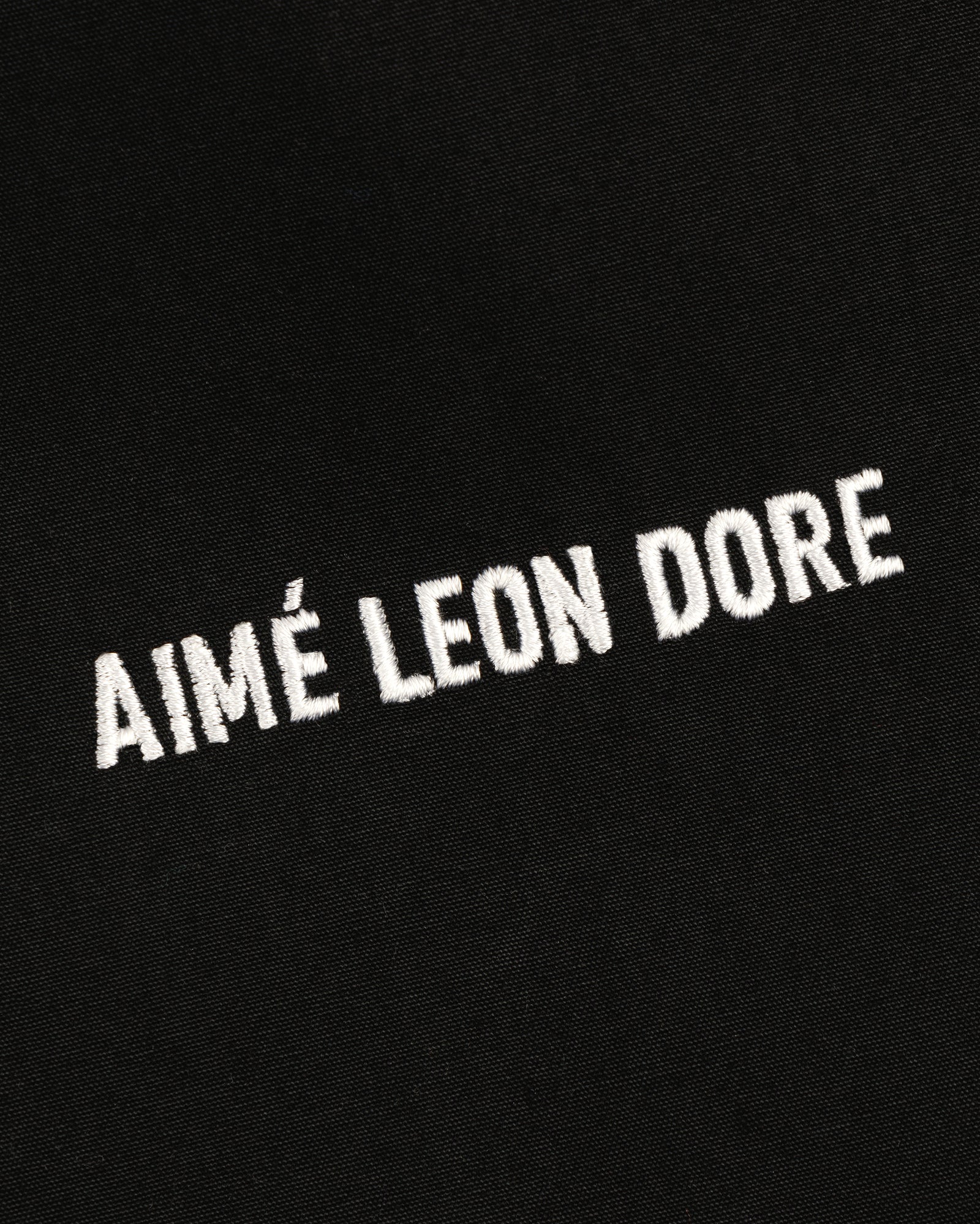 White “AIMÉ LEON DORE” text is embroidered on black Micro Logo Lightweight Jacket, angled upward left to right.