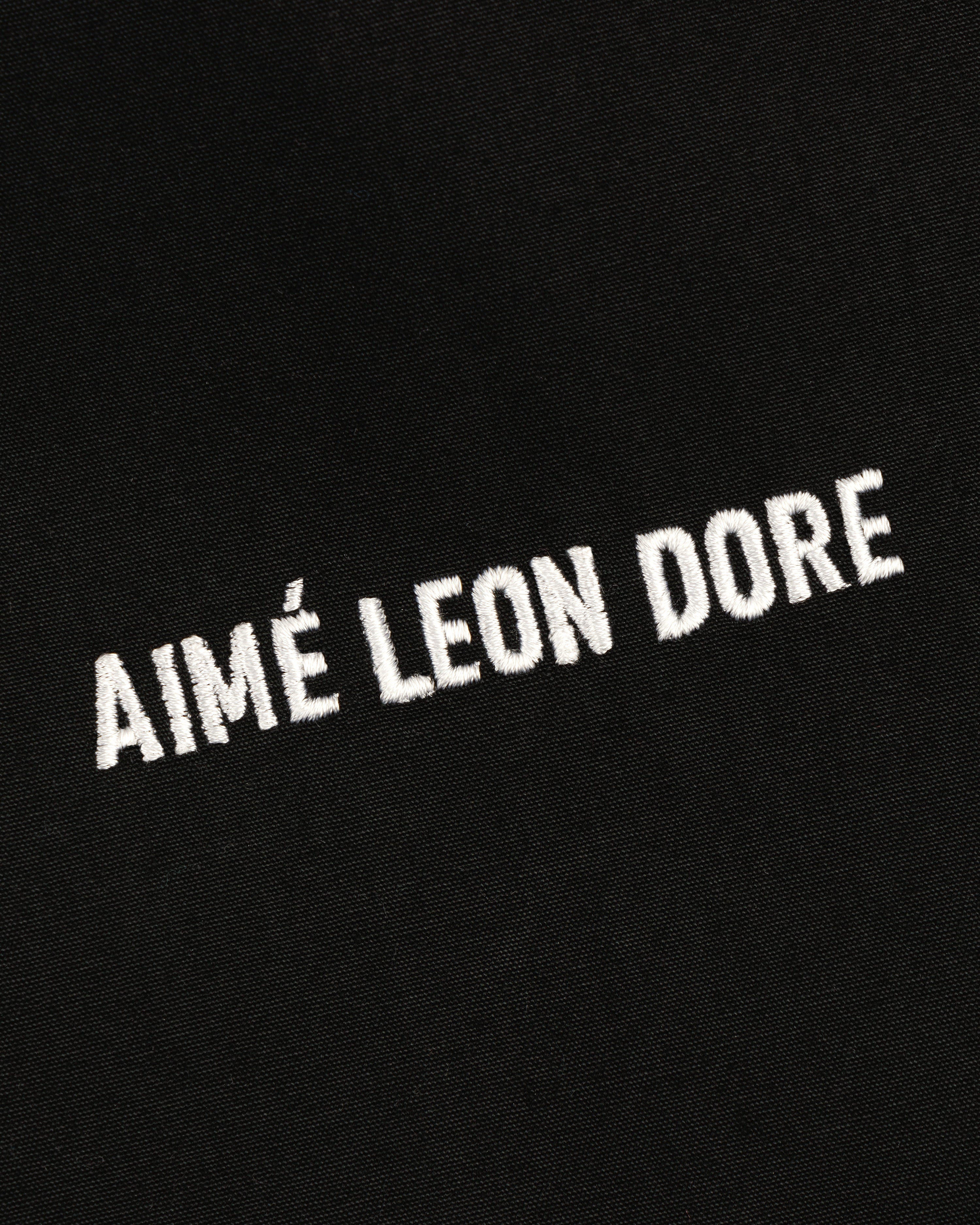 White “AIMÉ LEON DORE” text is embroidered on black Micro Logo Lightweight Jacket, angled upward left to right.