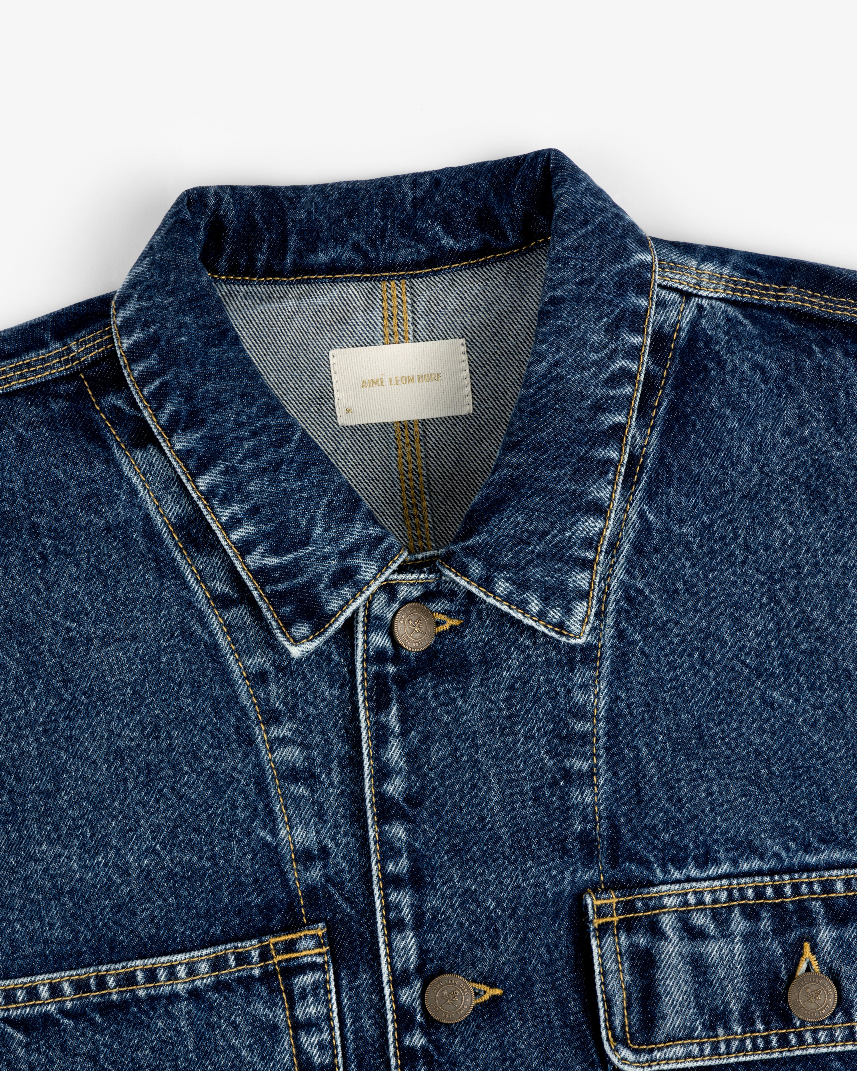 Close-up of the Denim Trucker Jacket in blue with visible stitching, metal buttons, and chest pockets.