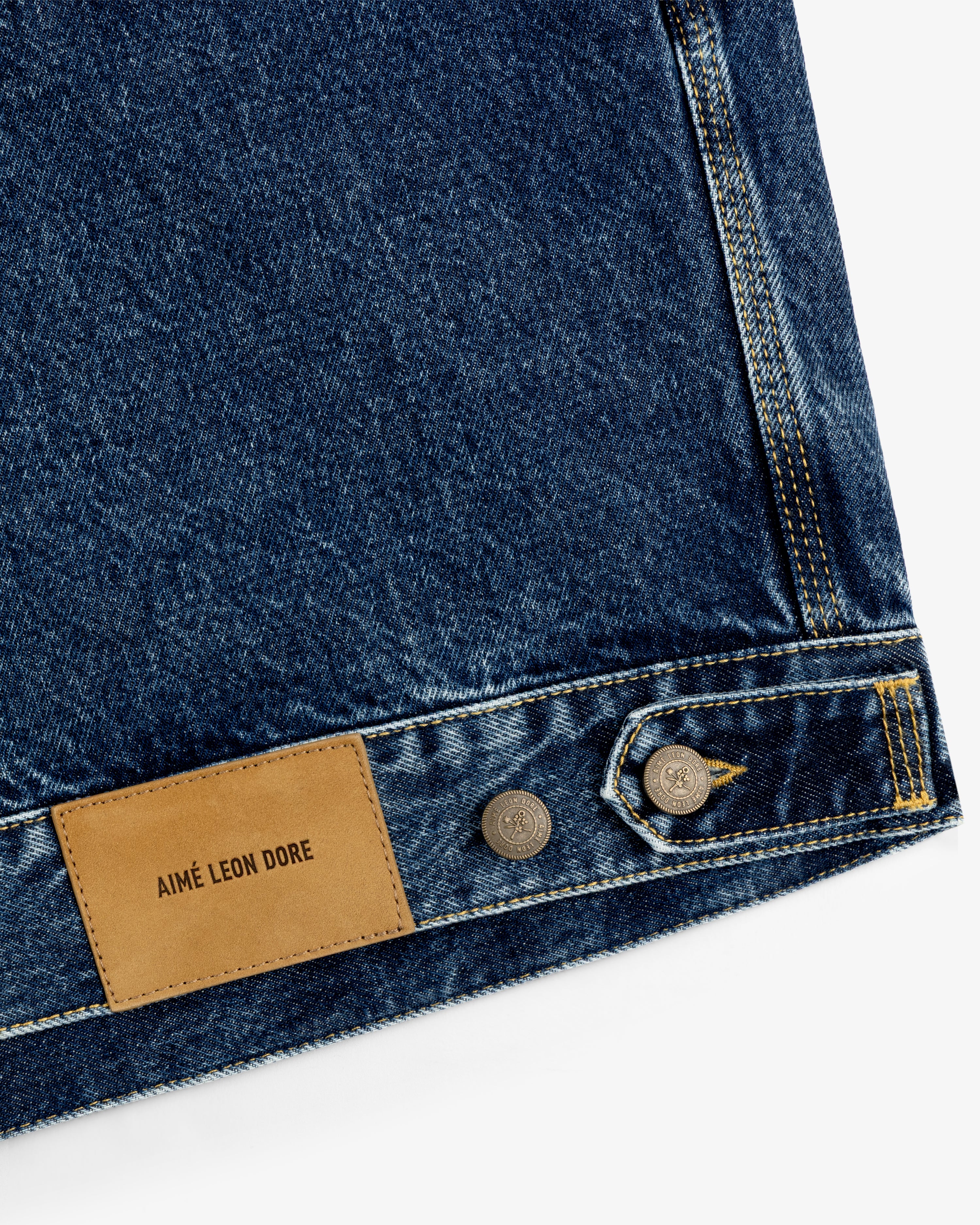 Close-up of blue denim Trucker Jacket with brown leather Aimé Leon Dore patch and silver buttons.