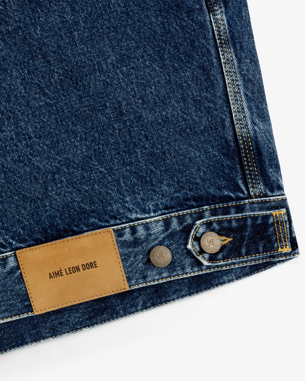Close-up of blue denim Trucker Jacket with brown leather Aimé Leon Dore patch and silver buttons.