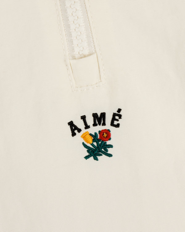 Close-up of the Crest Nylon Half-Zip Pullover with AIMÉ text above yellow and red embroidered flowers on cream fabric.