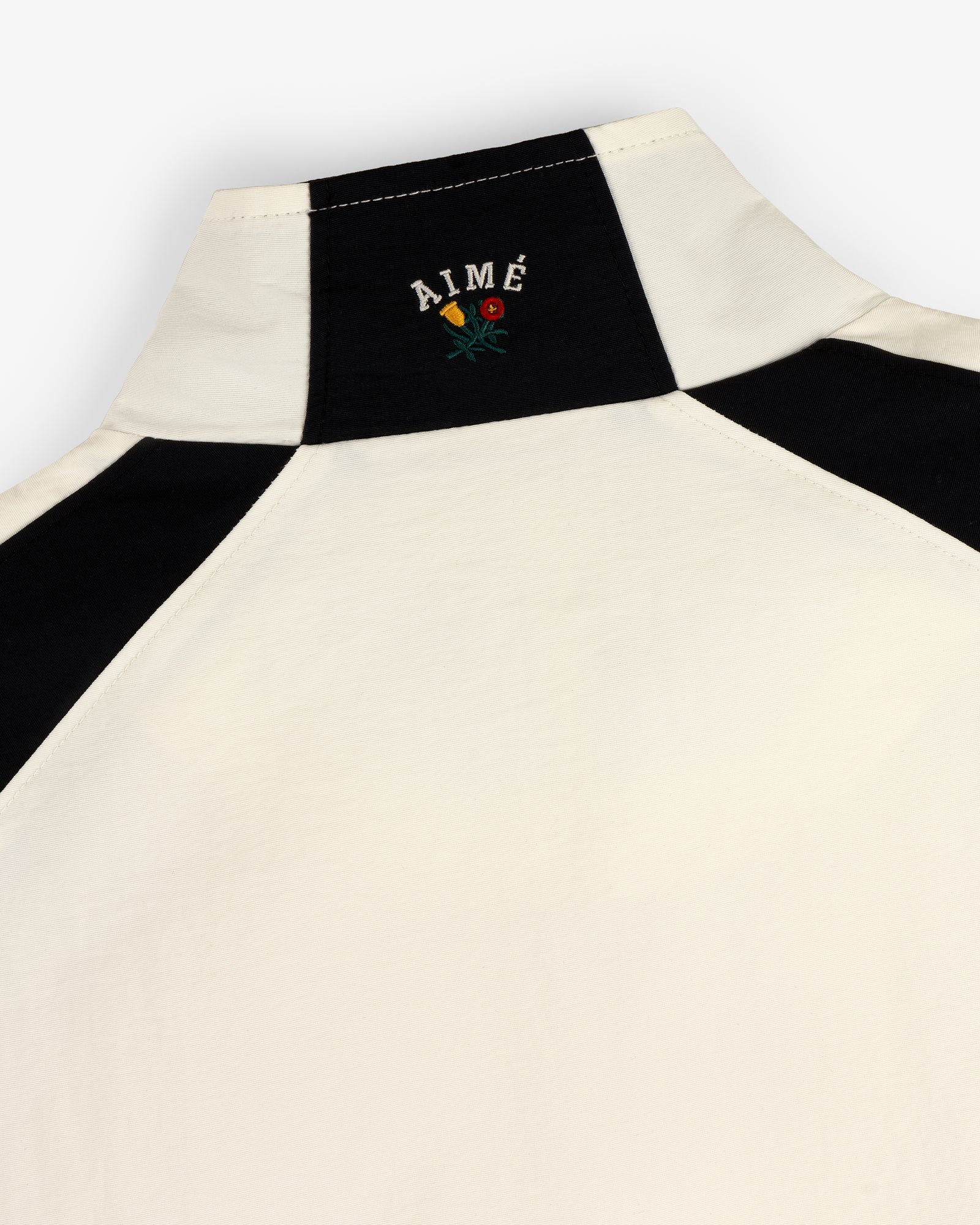 Close-up of a cream and black Crest Nylon Half-Zip Pullover with AIMÉ and a small embroidered flower on the back neck.