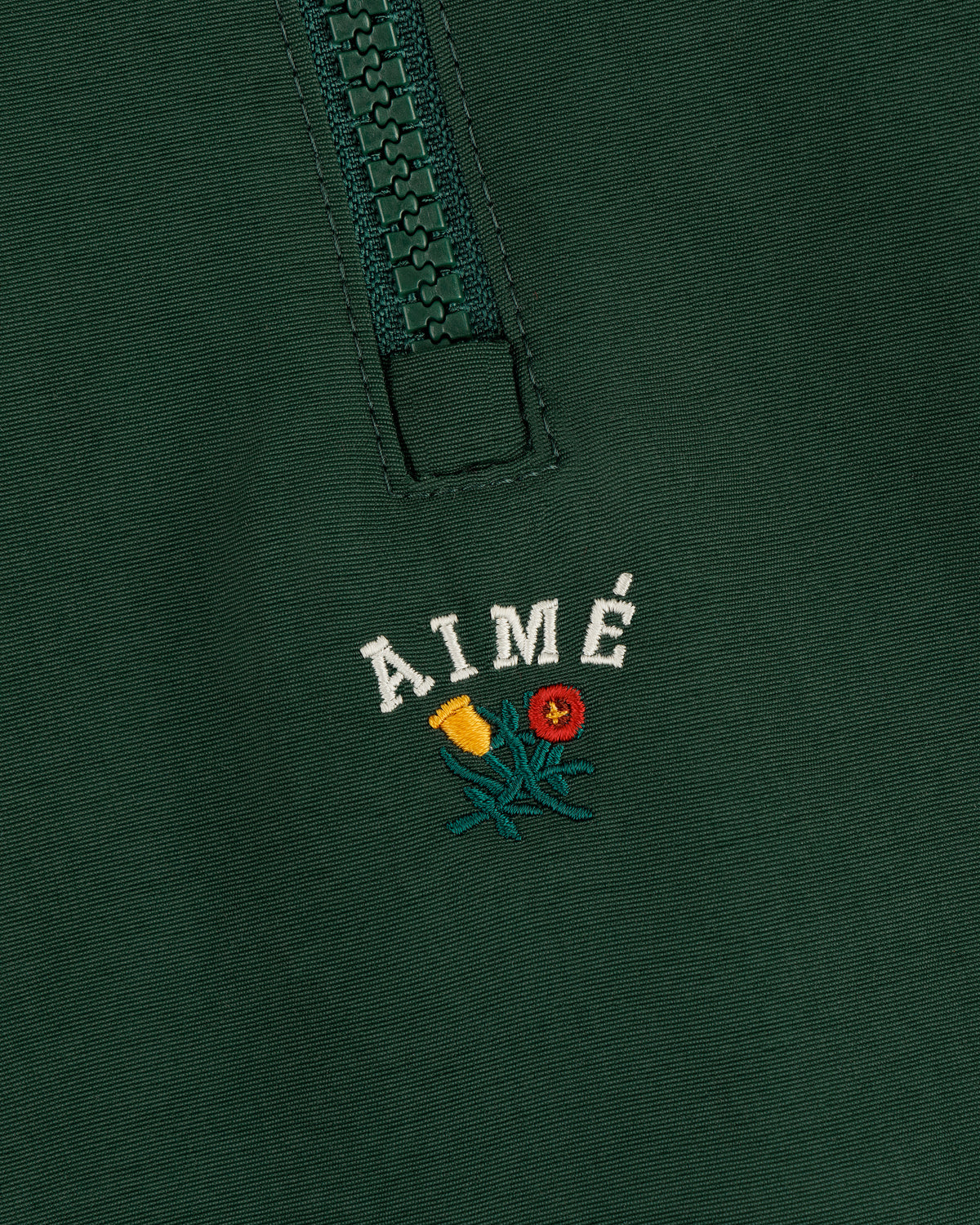 Close-up of a green Crest Nylon Half-Zip Pullover with AIMÉ embroidered above two small flowers.