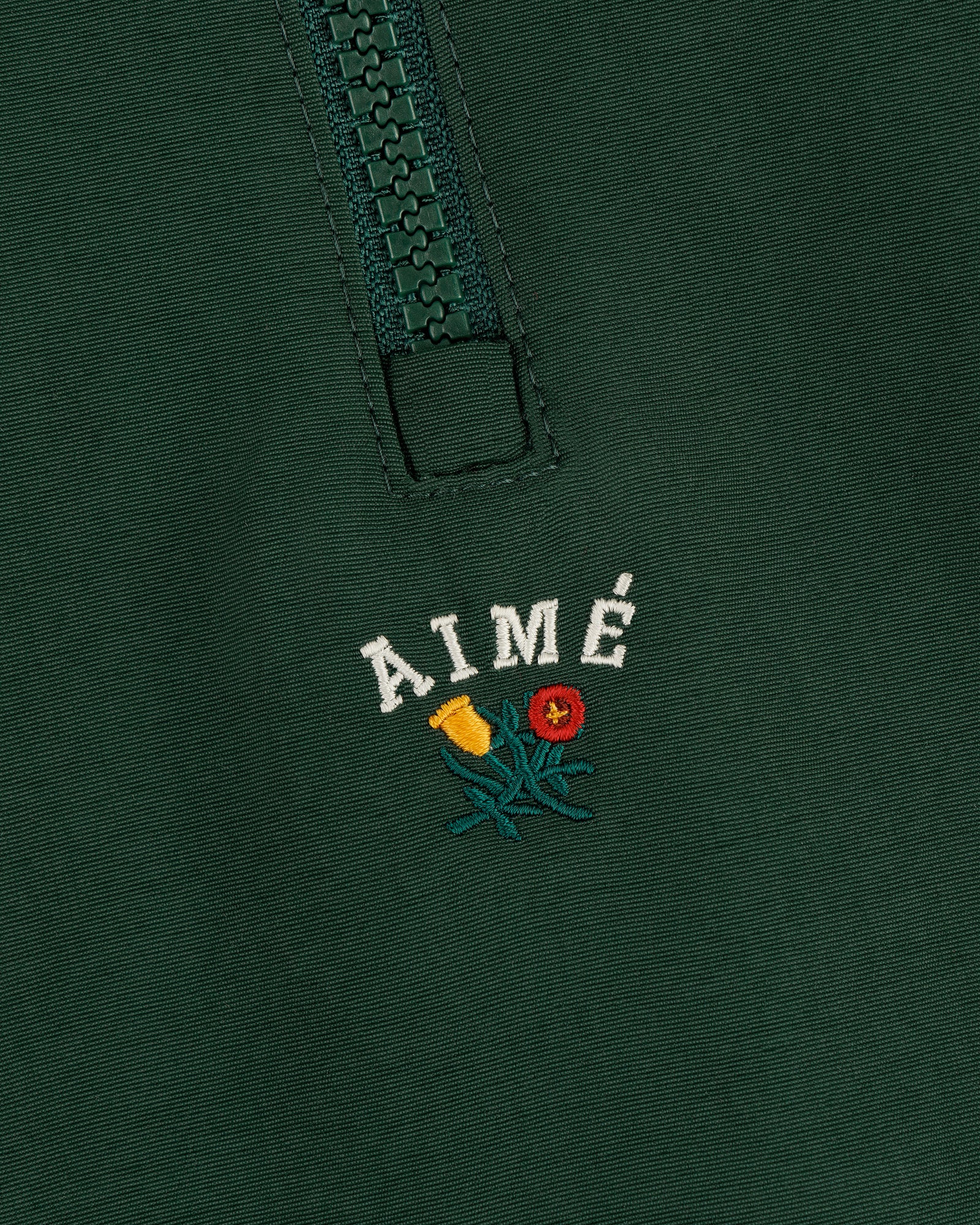 Close-up of a green Crest Nylon Half-Zip Pullover with AIMÉ embroidered above two small flowers.