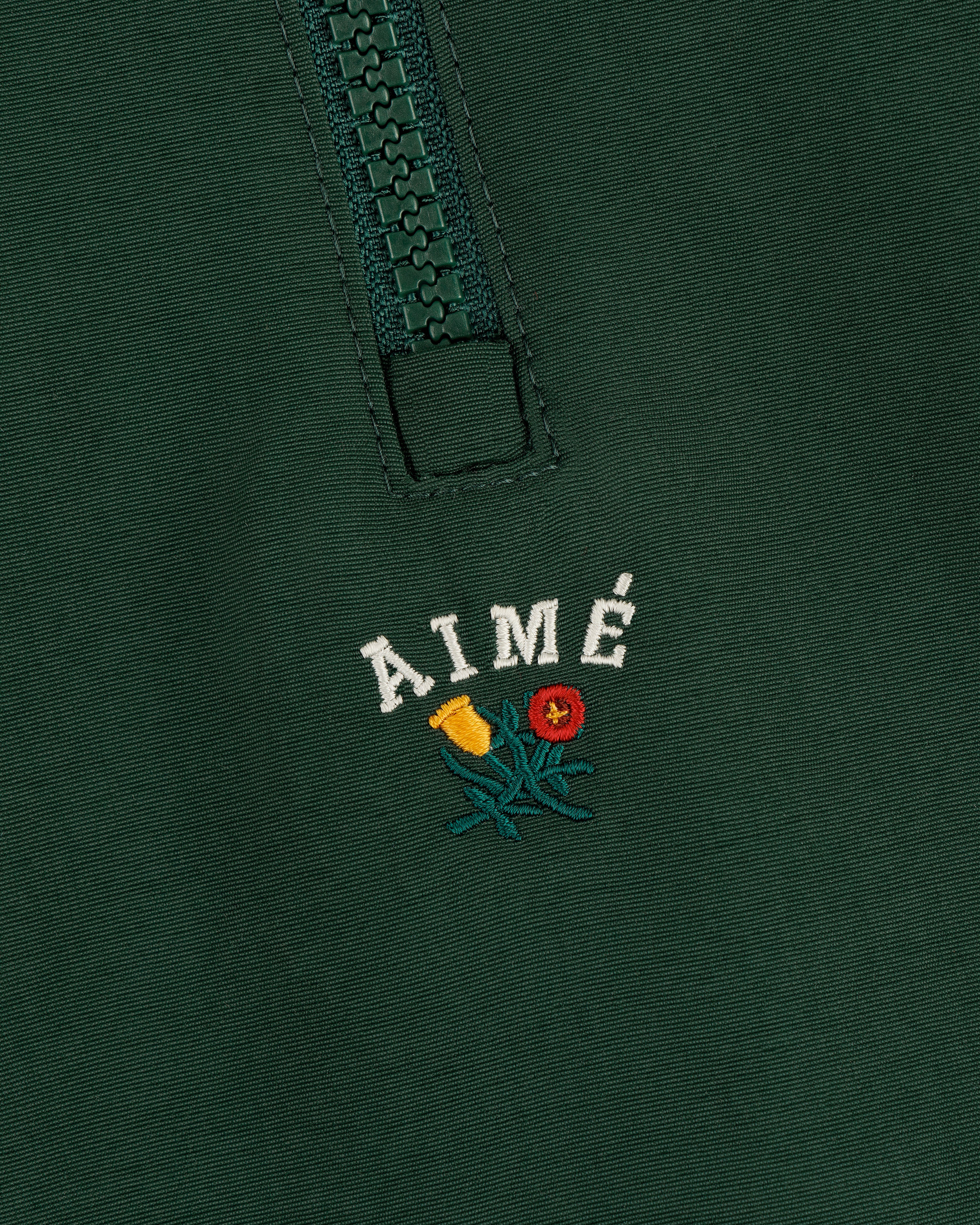 Close-up of a green Crest Nylon Half-Zip Pullover with AIMÉ embroidered above two small flowers.