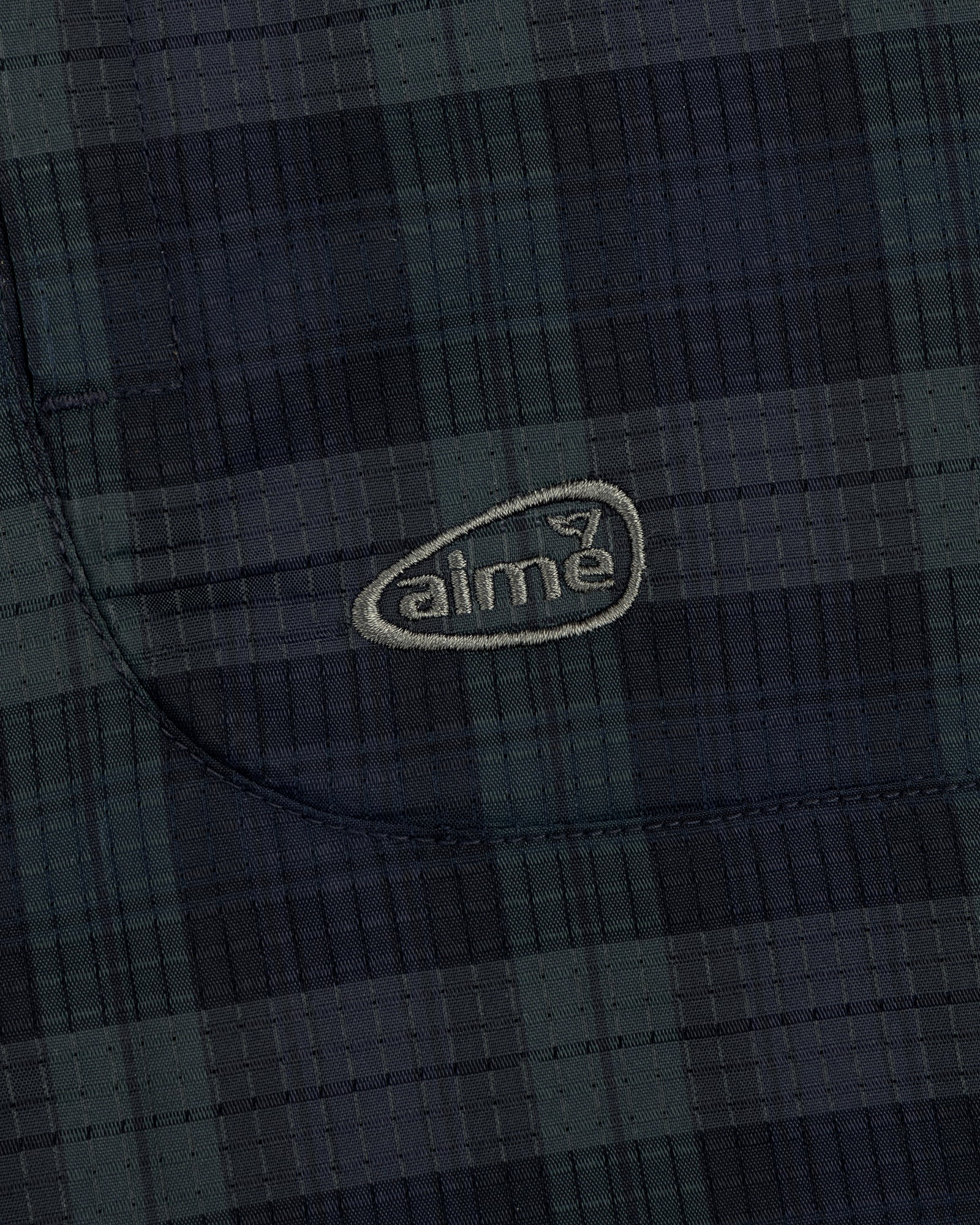 Close-up of ALD Golf Lightweight Tips Windbreaker showing dark plaid fabric, stitched pocket, and light aime logo.