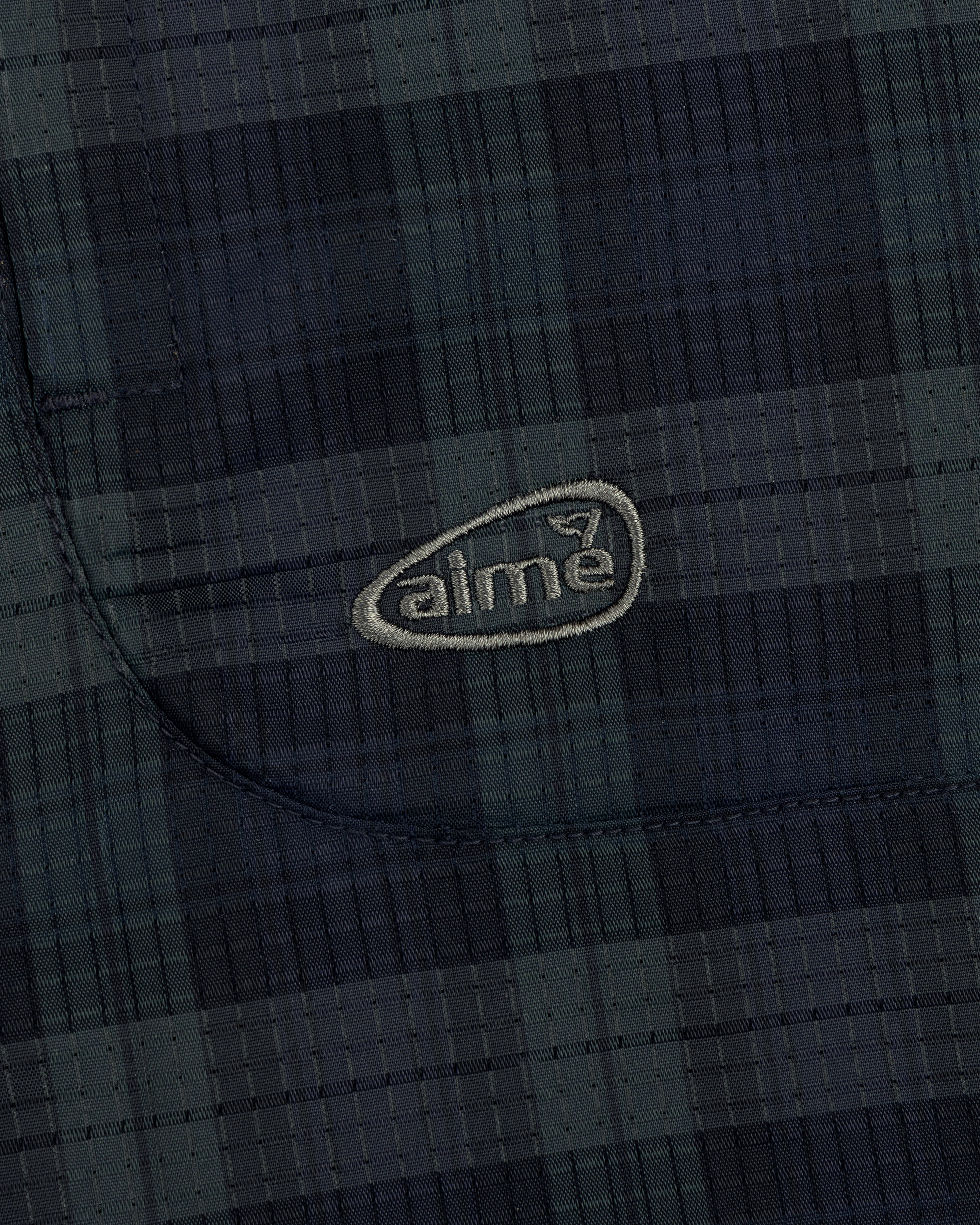 Close-up of ALD Golf Lightweight Tips Windbreaker showing dark plaid fabric, stitched pocket, and light aime logo.