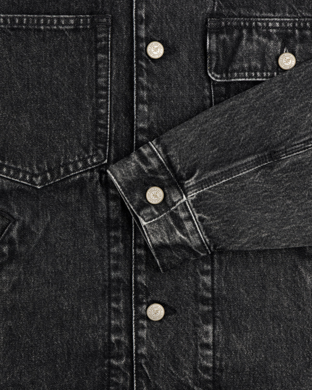 Close-up of a Denim Trucker Jacket in black with silver buttons, chest pockets, and one sleeve across the front.