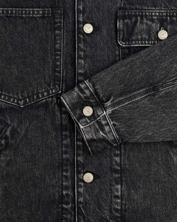 Close-up of a Denim Trucker Jacket in black with silver buttons, chest pockets, and one sleeve across the front.
