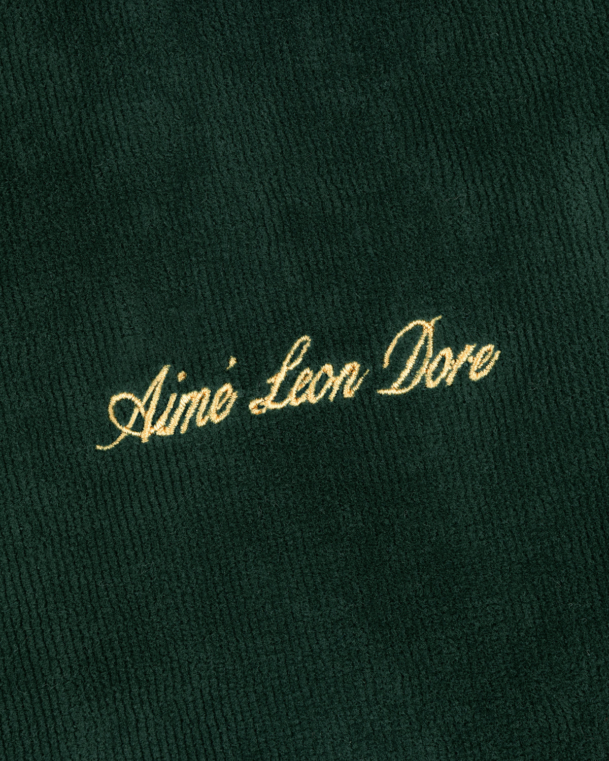 Gold script text Aimé Leon Dore embroidered on dark green ribbed Mulberry Robe fabric.