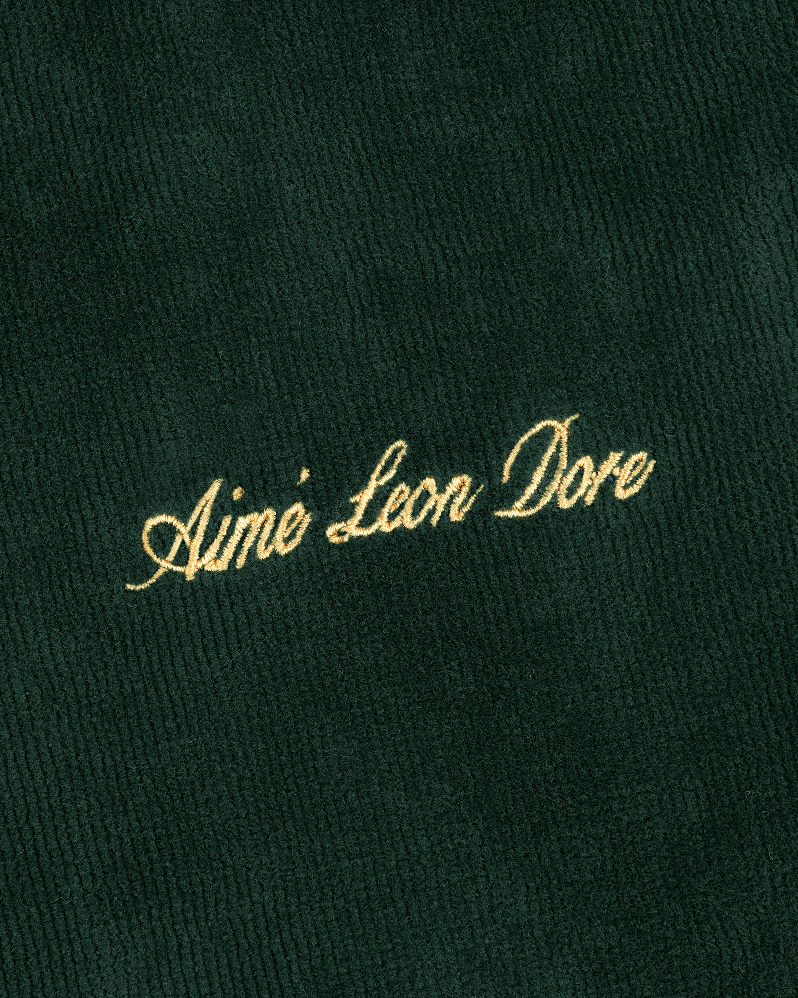 Gold script text Aimé Leon Dore embroidered on dark green ribbed Mulberry Robe fabric.