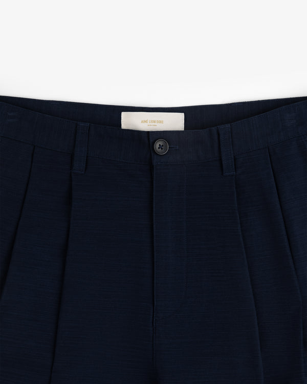 Double Pleated Pant: Navy blue dress pants with waistband, belt loops, pleats, and a black button above the zipper.