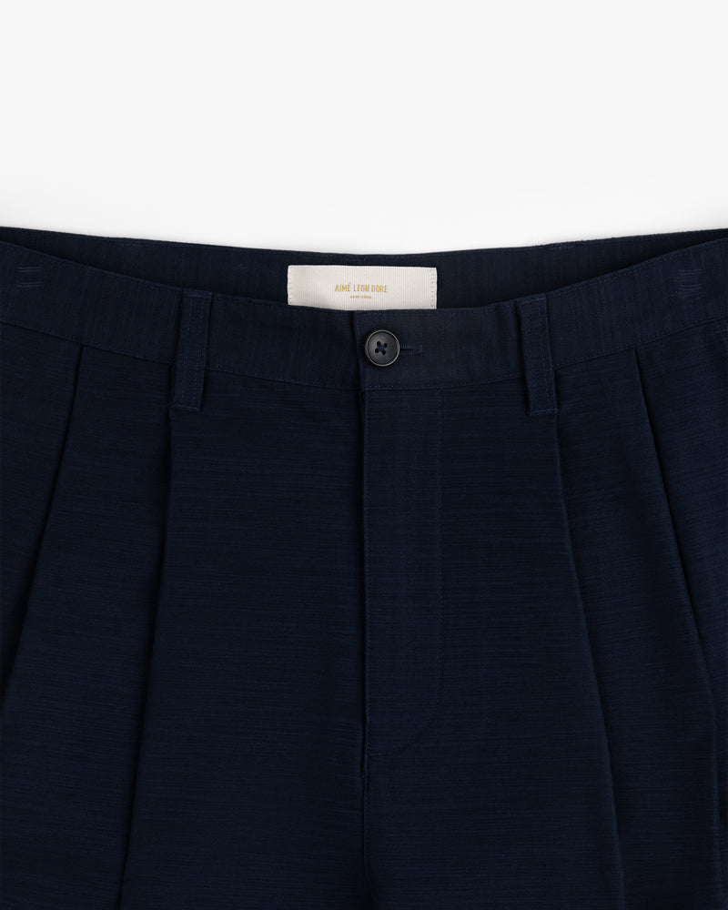 Double Pleated Pant: Navy blue dress pants with waistband, belt loops, pleats, and a black button above the zipper.