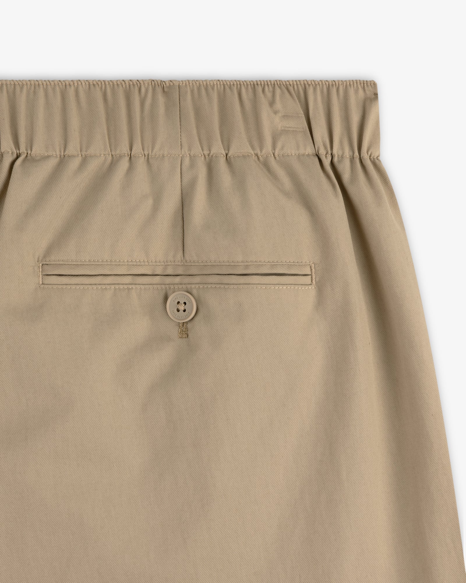 Relaxed Cotton Trouser in tan with elastic waistband and single buttoned back pocket, shown on a white background.