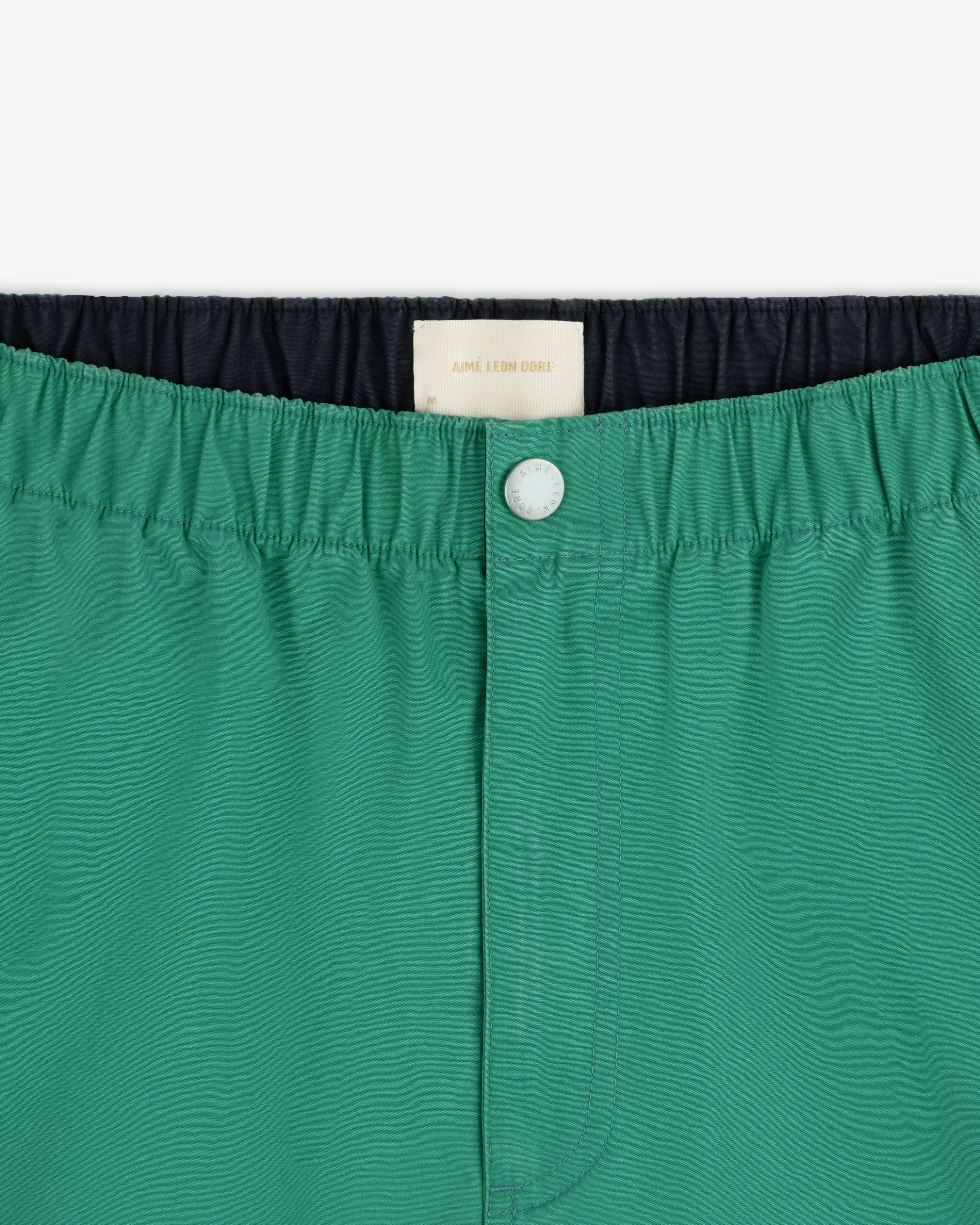 Close-up of Washed Cotton Windbreaker Pant in green, featuring an elastic waistband, front button, and white inner label.