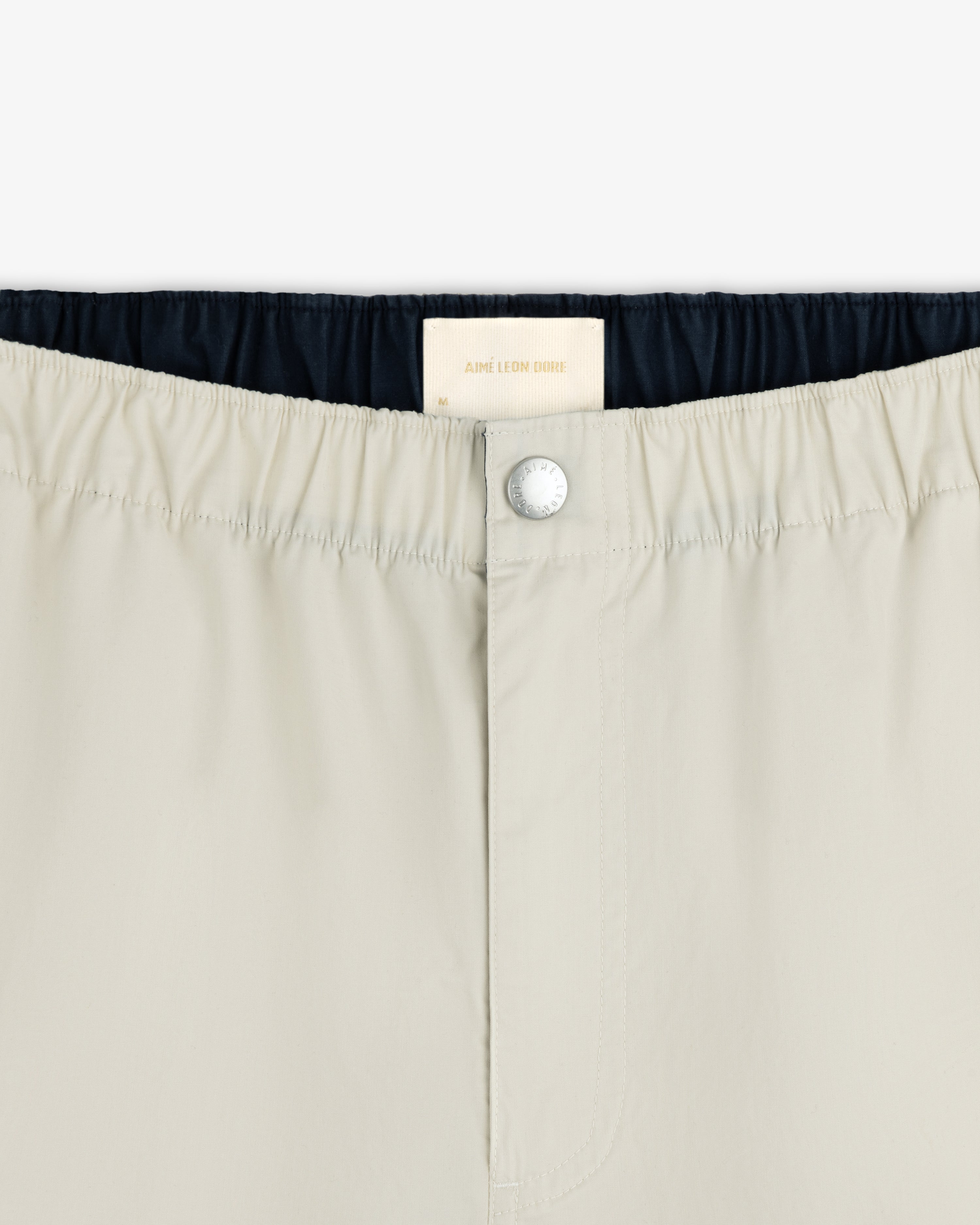 Close-up of Washed Cotton Windbreaker Pant in beige with elastic waistband, button closure, and Aimé Leon Dore label.