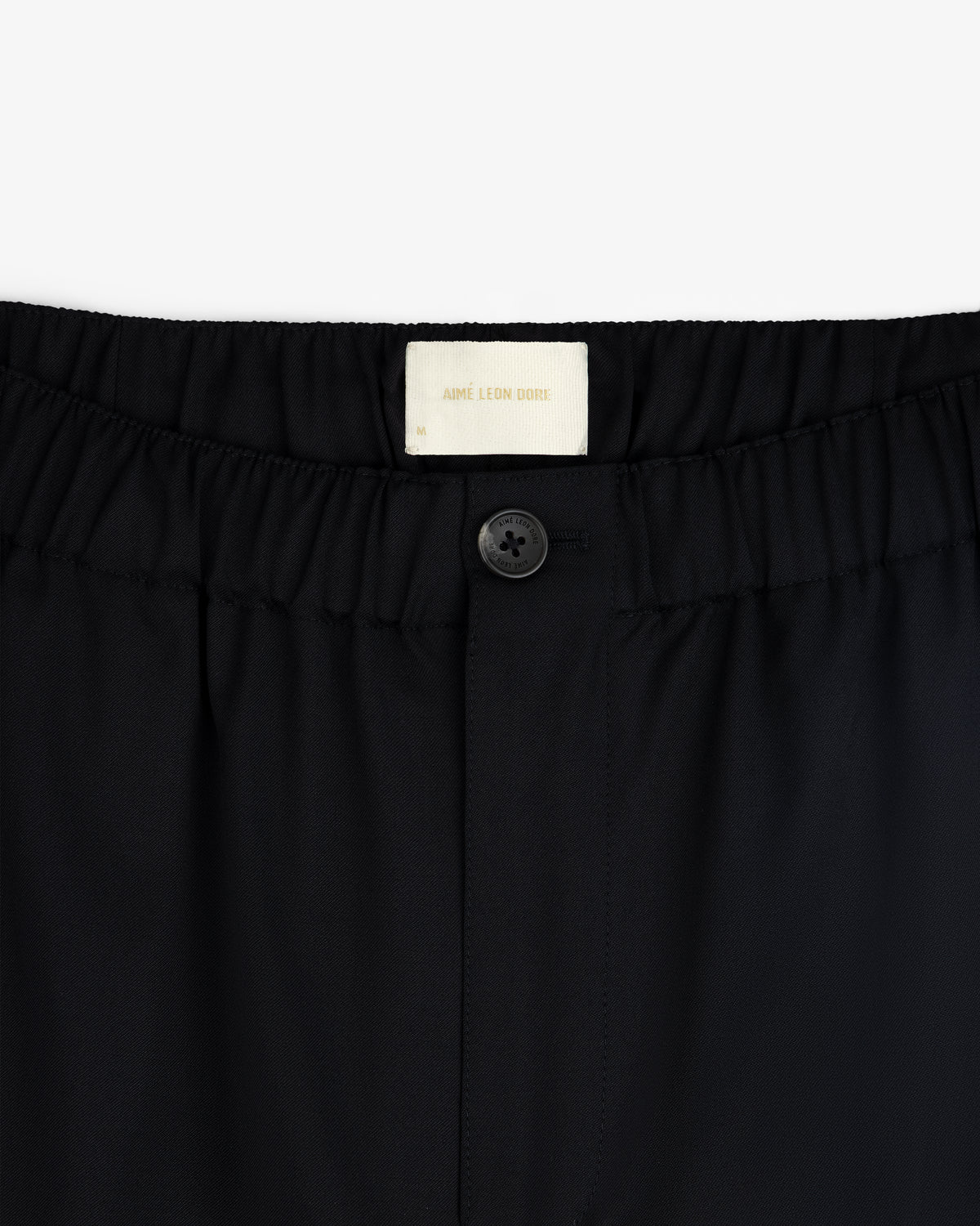 Close-up of Elasticated Waist Gabardine Trouser in black with a button closure and visible Aimé Leon Dore label.