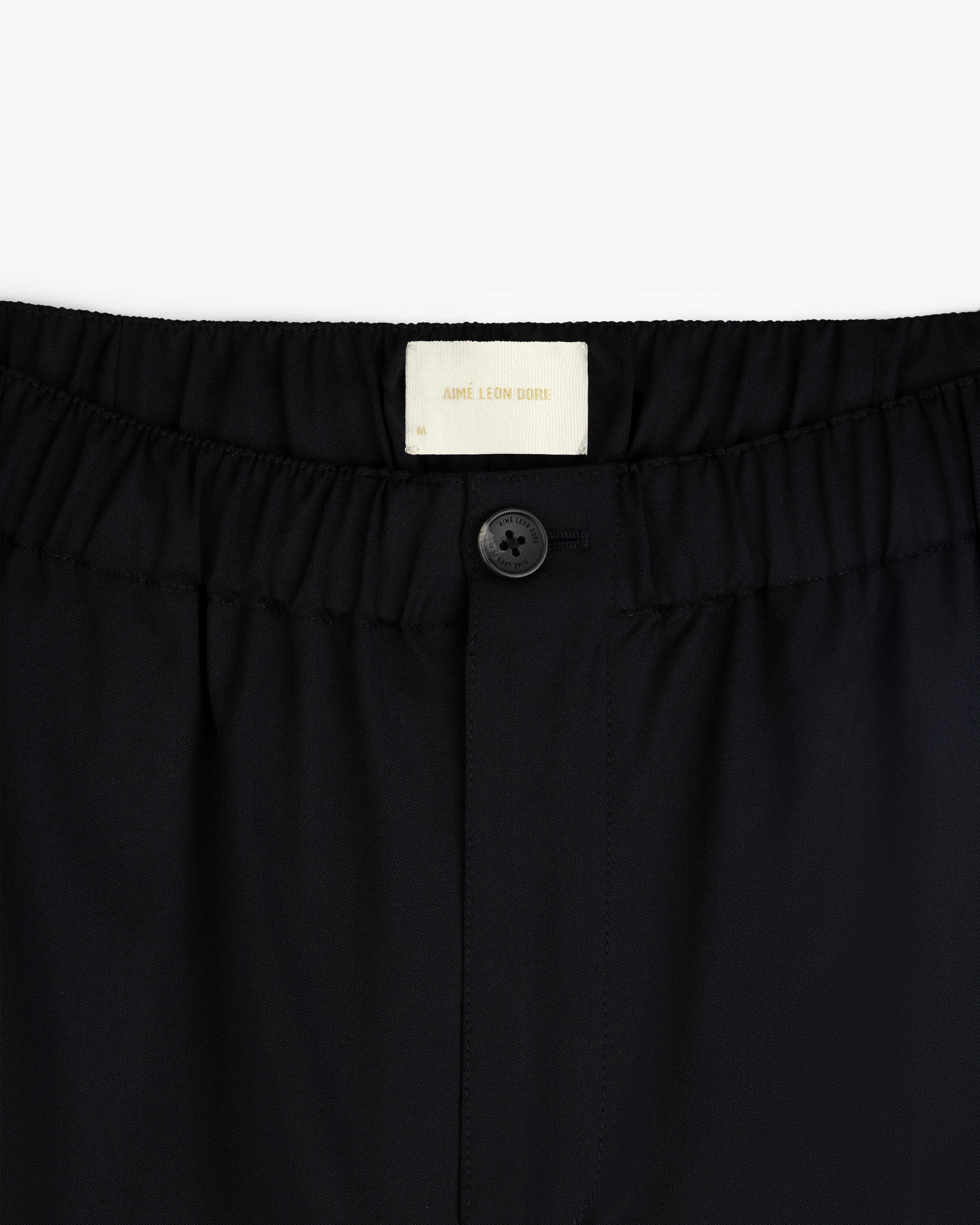 Close-up of Elasticated Waist Gabardine Trouser in black with a button closure and visible Aimé Leon Dore label.