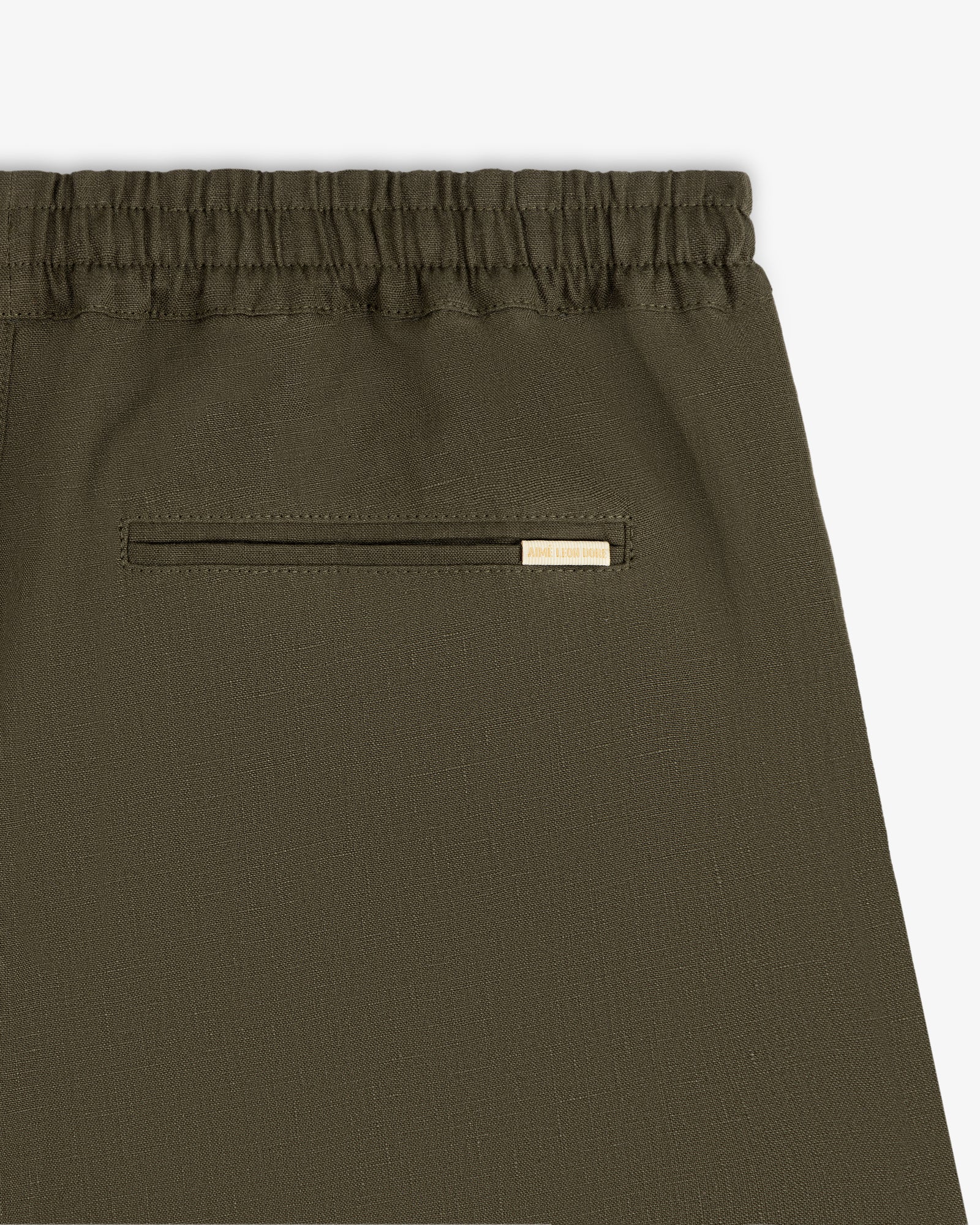 Close-up of Linen Leisure Pant in olive green with elastic waistband and a small pocket topped by a gold tag.