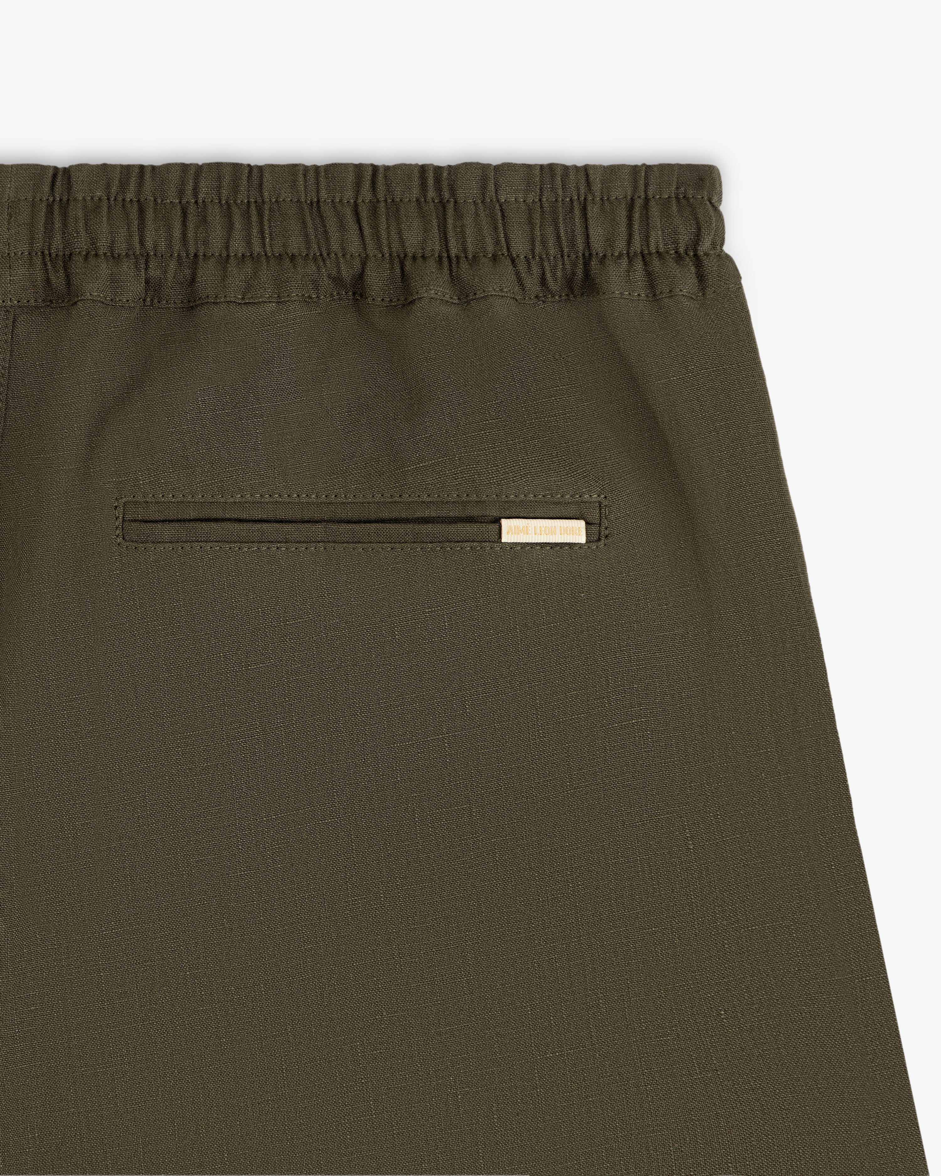Close-up of Linen Leisure Pant in olive green with elastic waistband and a small pocket topped by a gold tag.
