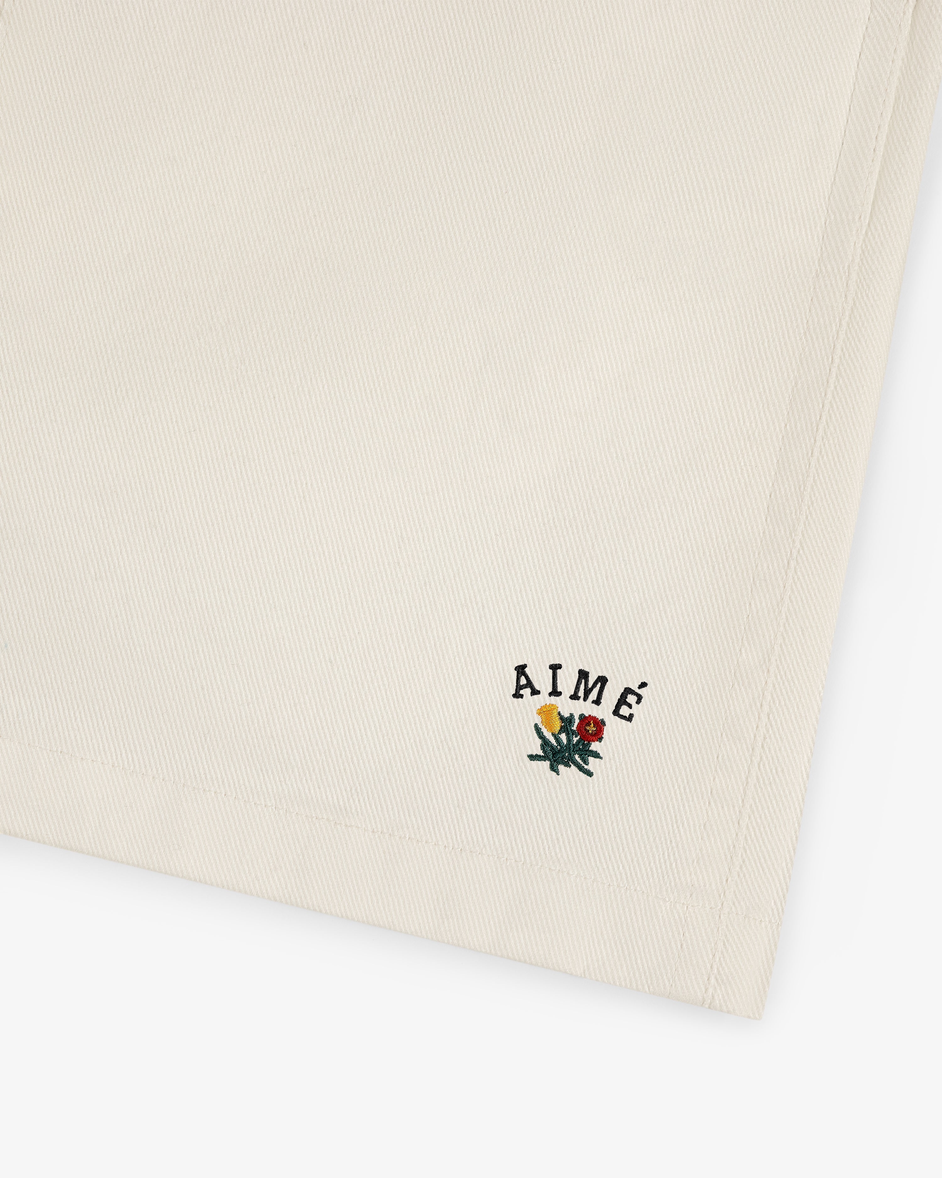 Beige Crest Casual Short with small embroidered AIMÉ text and a floral design in the bottom corner.