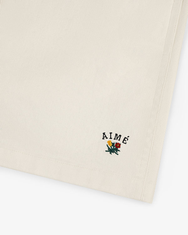 Beige Crest Casual Short with small embroidered AIMÉ text and a floral design in the bottom corner.