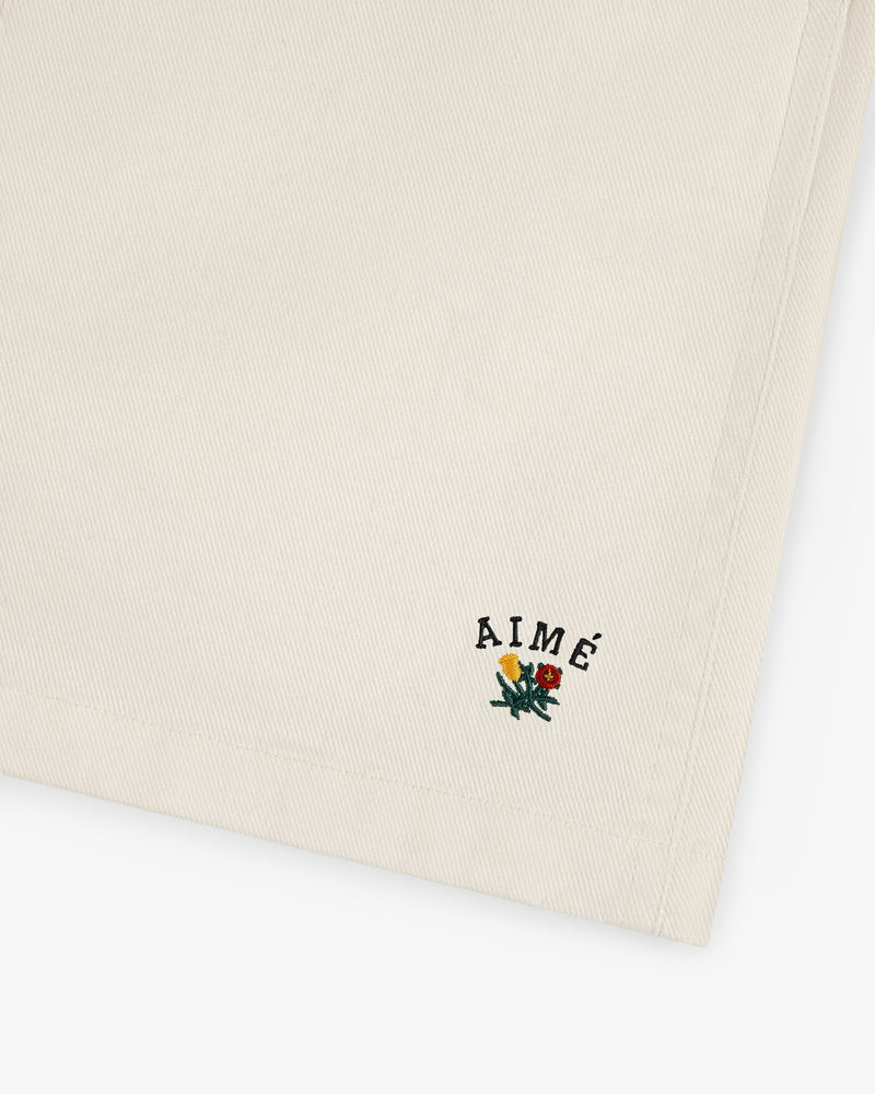 Beige Crest Casual Short with small embroidered AIMÉ text and a floral design in the bottom corner.