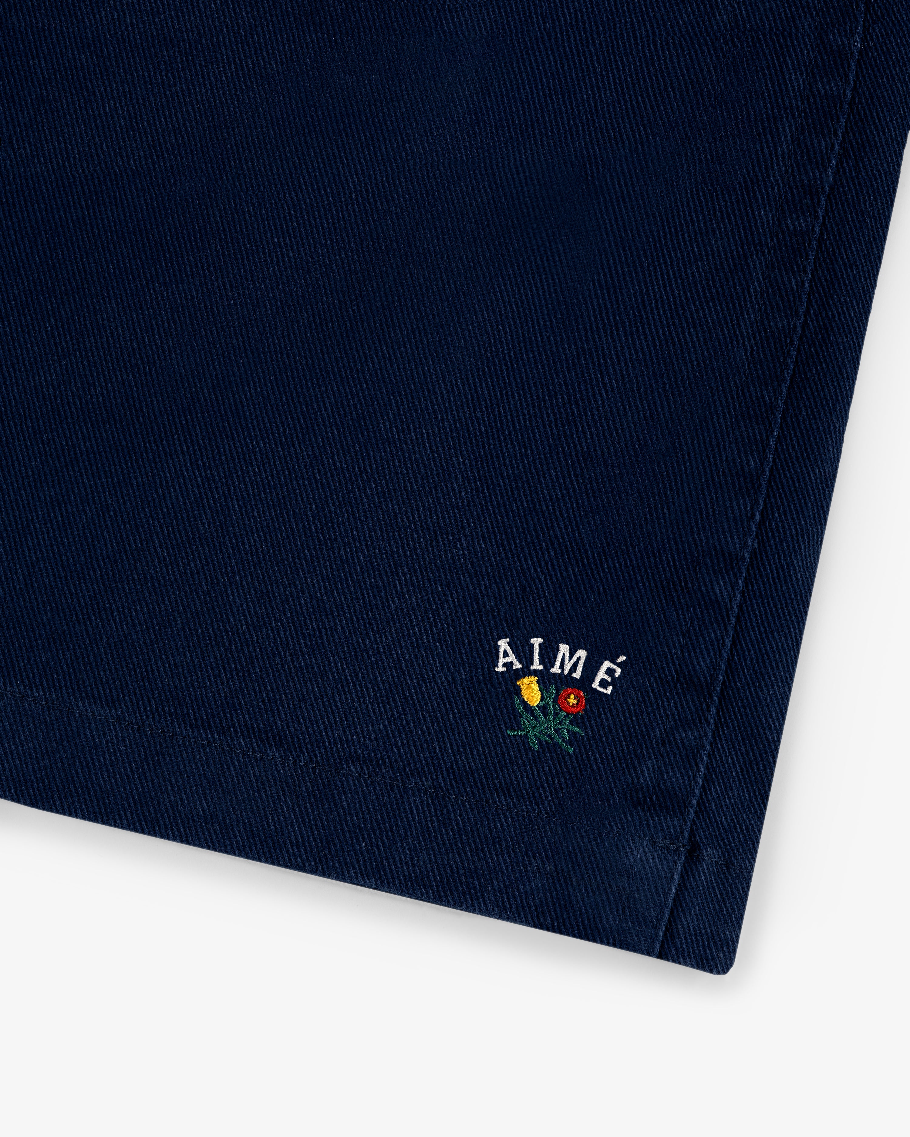 Close-up of Crest Casual Short in navy with AIMÉ and three small embroidered flowers near the hem.
