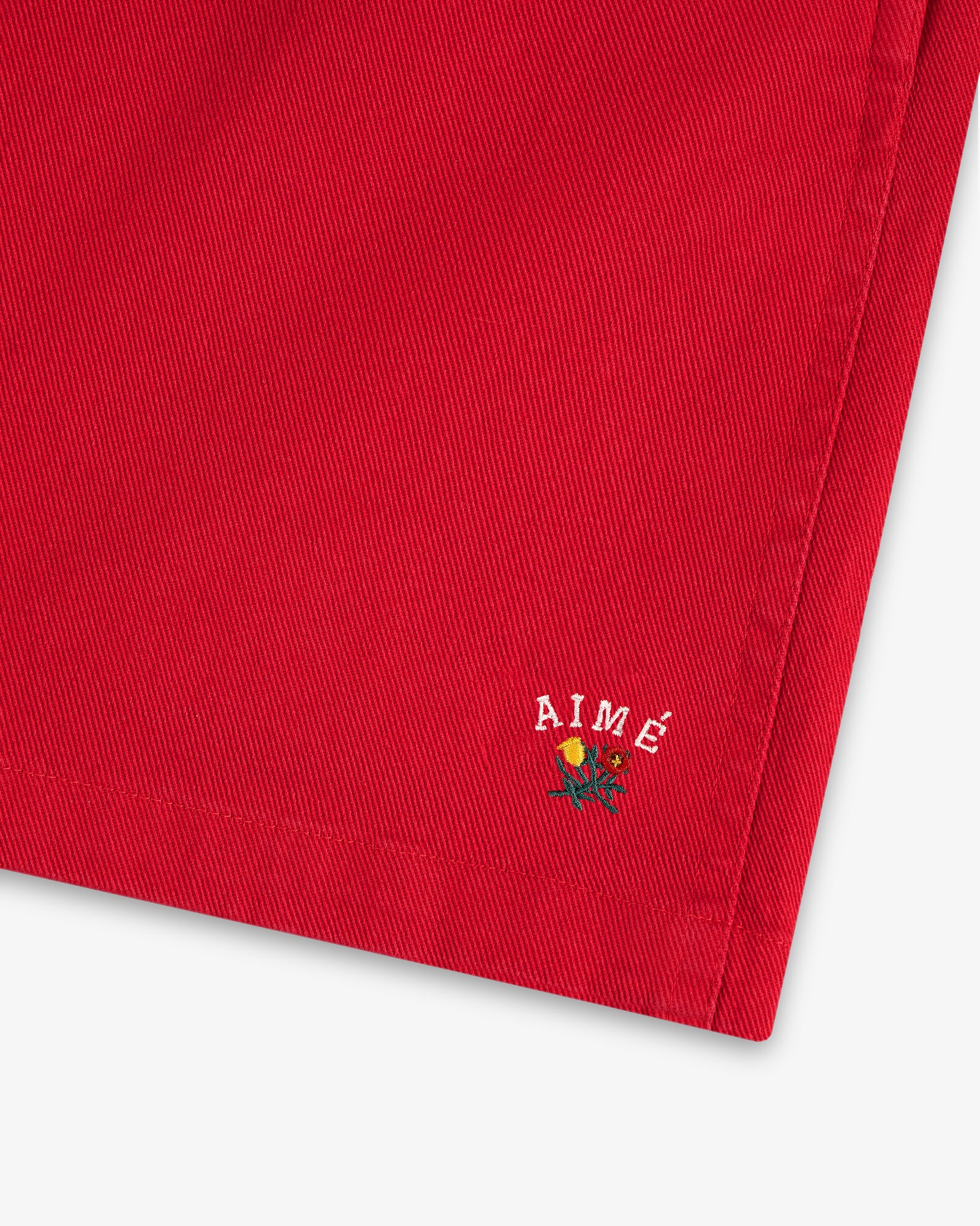 Close-up of Crest Casual Short's red fabric with small AIMÉ text and an embroidered yellow rose near the hem.