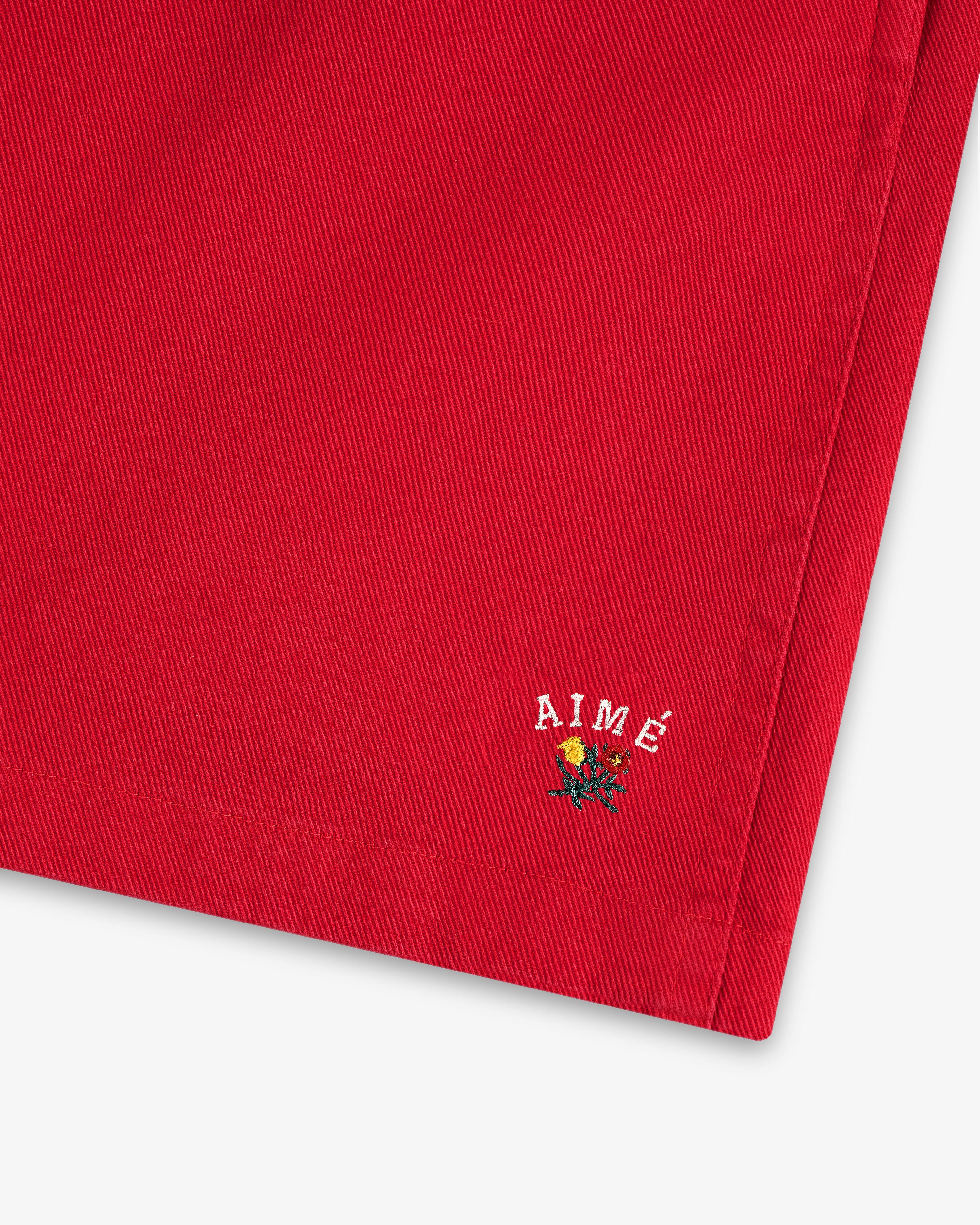Close-up of Crest Casual Short's red fabric with small AIMÉ text and an embroidered yellow rose near the hem.