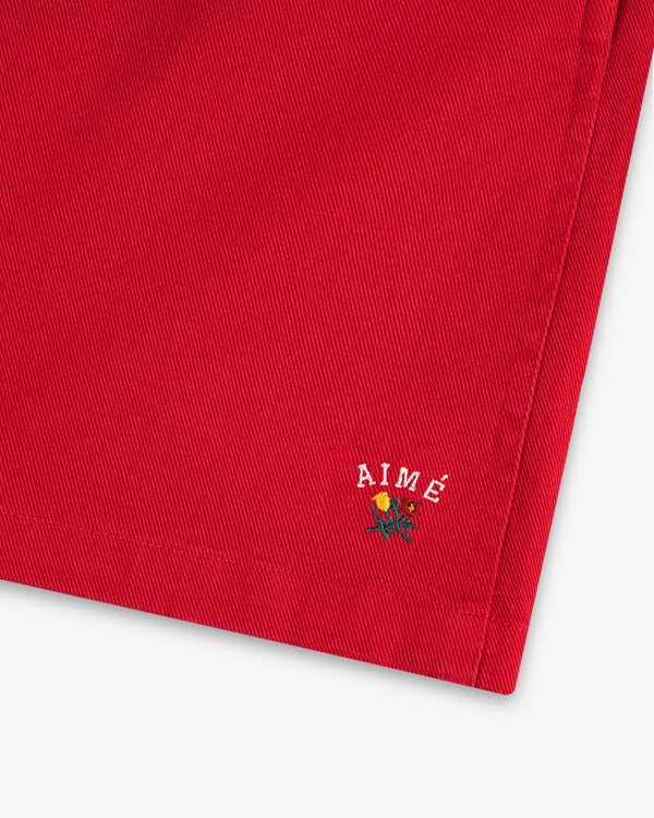 Close-up of Crest Casual Short's red fabric with small AIMÉ text and an embroidered yellow rose near the hem.