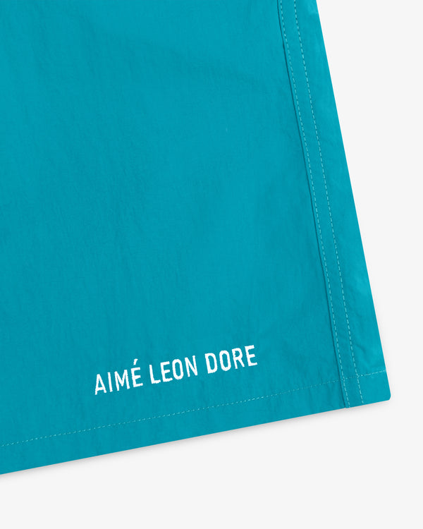 Close-up of turquoise Logo Swim Short with white AIMÉ LEON DORE text embroidered near the hem.