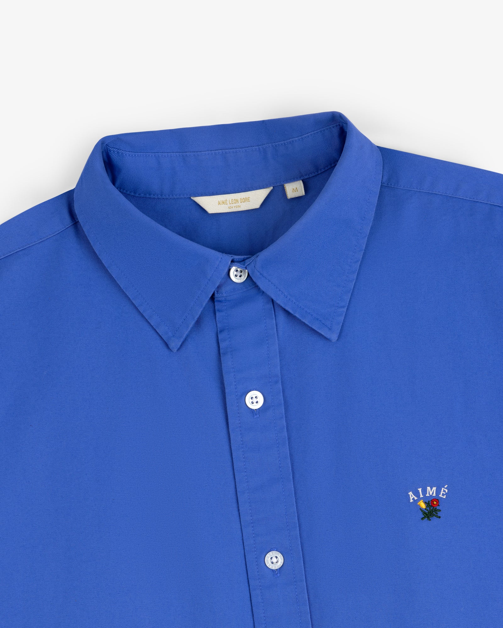 Leon Overshirt: Bright blue button-up with a collar and AIMÉ embroidered above a small rose on the chest.