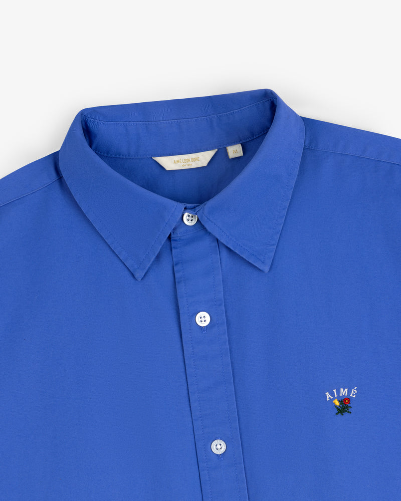 Leon Overshirt: Bright blue button-up with a collar and AIMÉ embroidered above a small rose on the chest.