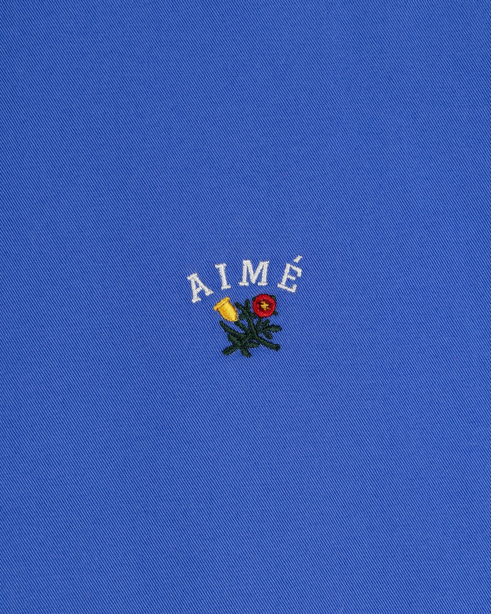 Leon Overshirt: blue fabric with AIMÉ embroidered in white above small yellow and red flowers with green leaves.