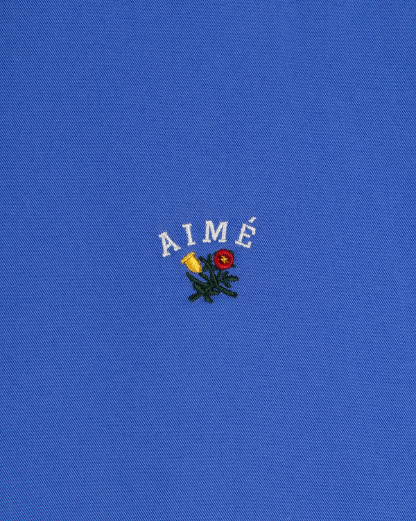 Leon Overshirt: blue fabric with AIMÉ embroidered in white above small yellow and red flowers with green leaves.