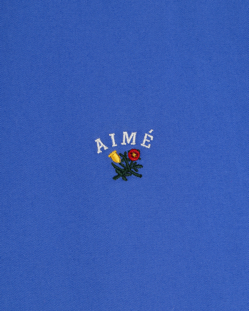 Leon Overshirt: blue fabric with AIMÉ embroidered in white above small yellow and red flowers with green leaves.