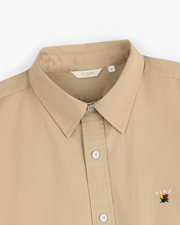 The Leon Overshirt is a beige collared button-up with a small embroidered AIMÉ logo and rose near the bottom right.