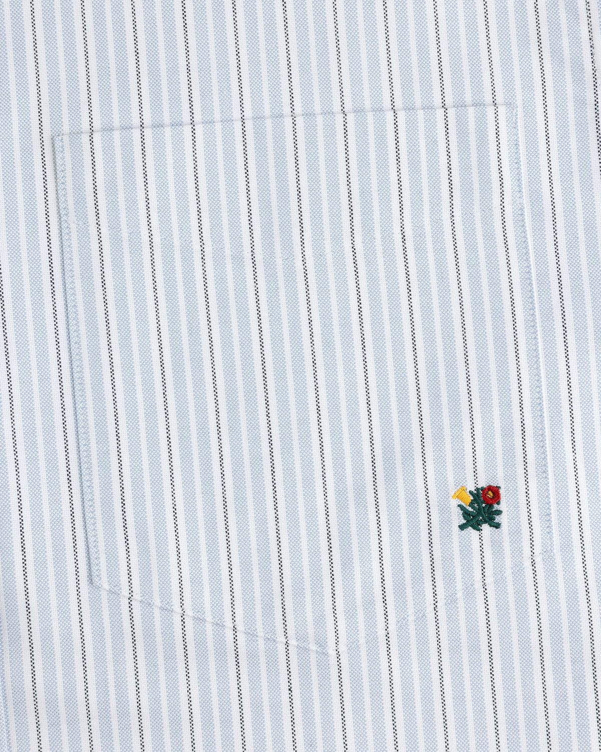 The Yarn Dyed Striped Crest Shirt is white with thin blue stripes and a small embroidered flower near the hem.
