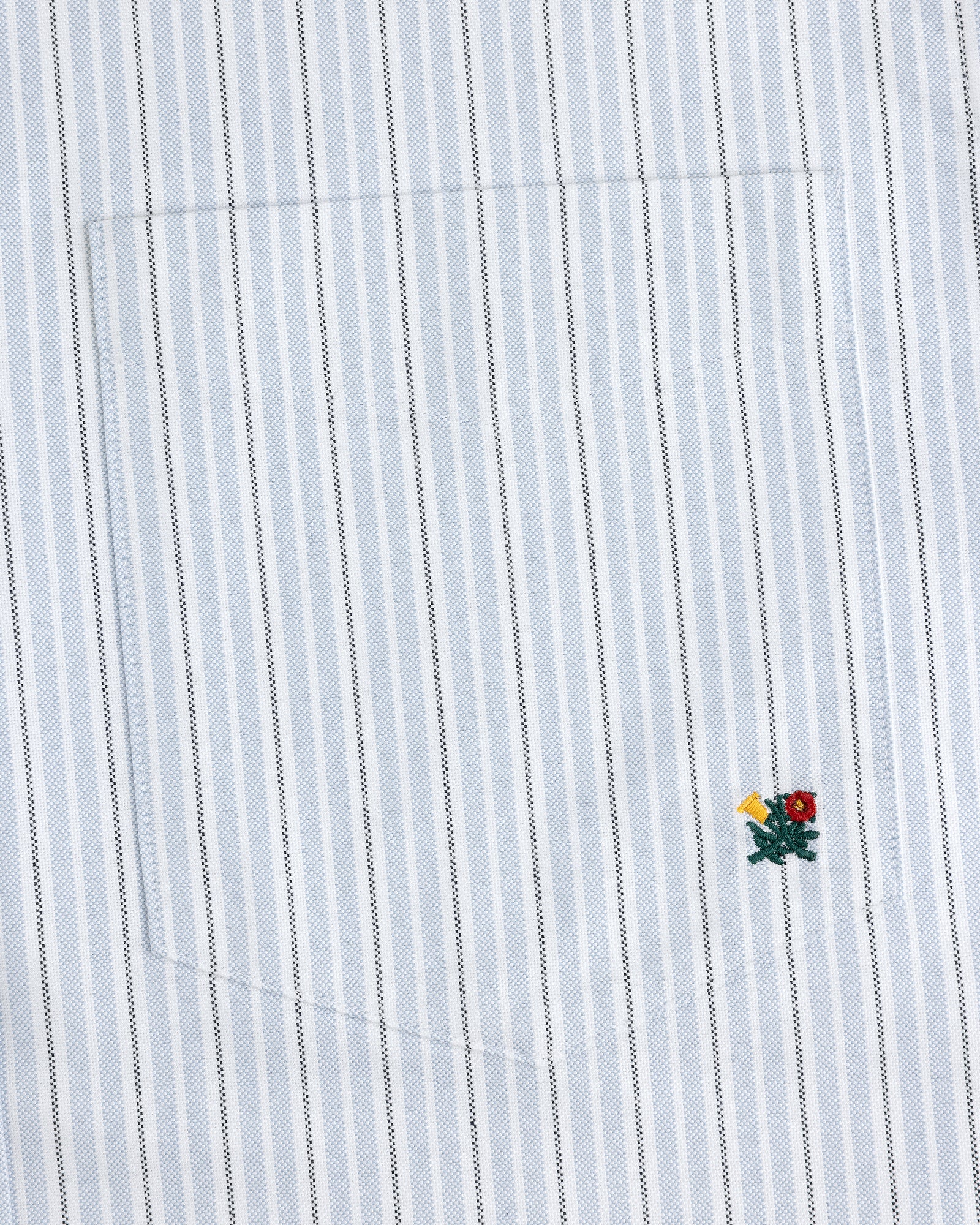 The Yarn Dyed Striped Crest Shirt is white with thin blue stripes and a small embroidered flower near the hem.