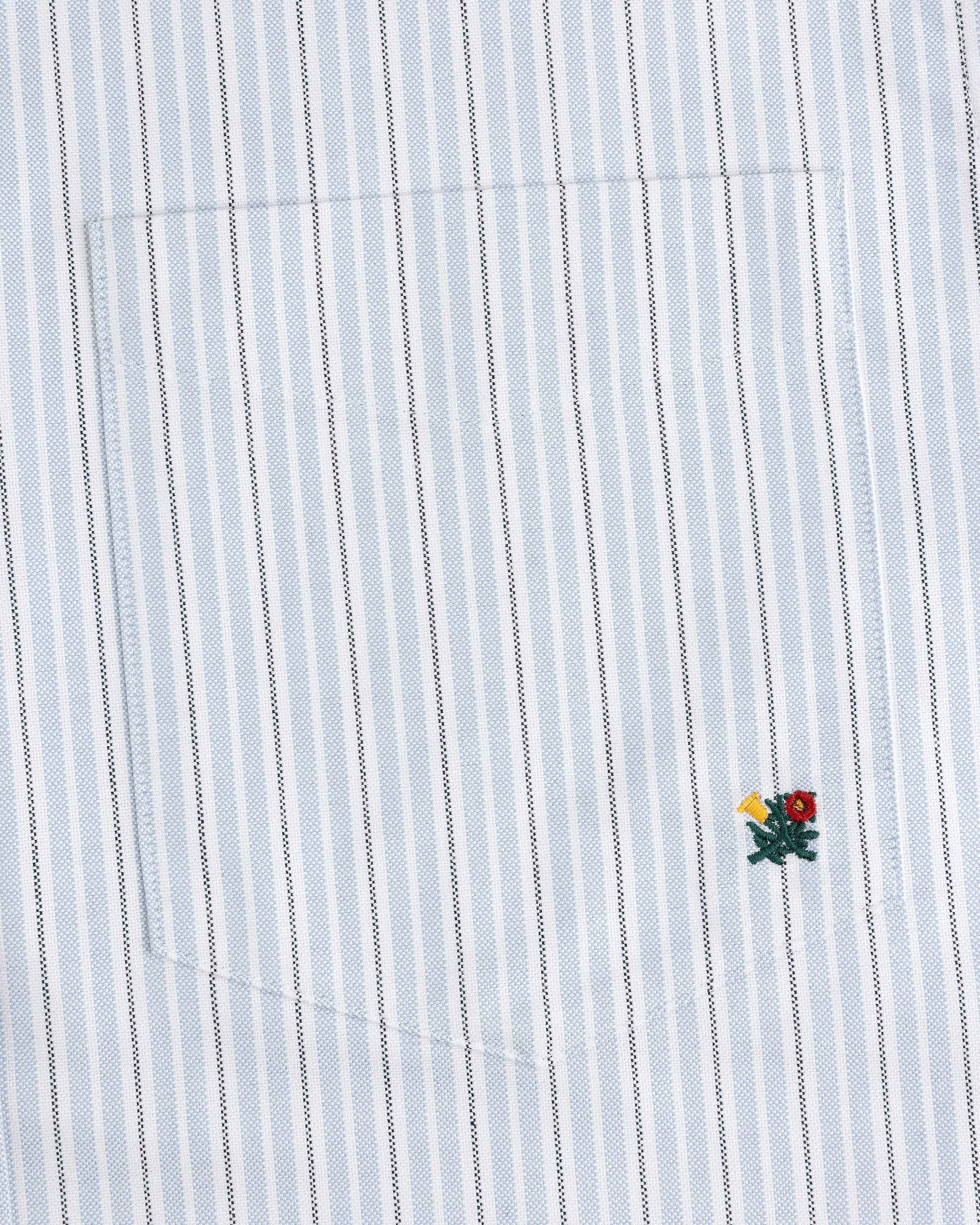 The Yarn Dyed Striped Crest Shirt is white with thin blue stripes and a small embroidered flower near the hem.