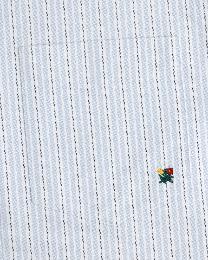 The Yarn Dyed Striped Crest Shirt is white with thin blue stripes and a small embroidered flower near the hem.