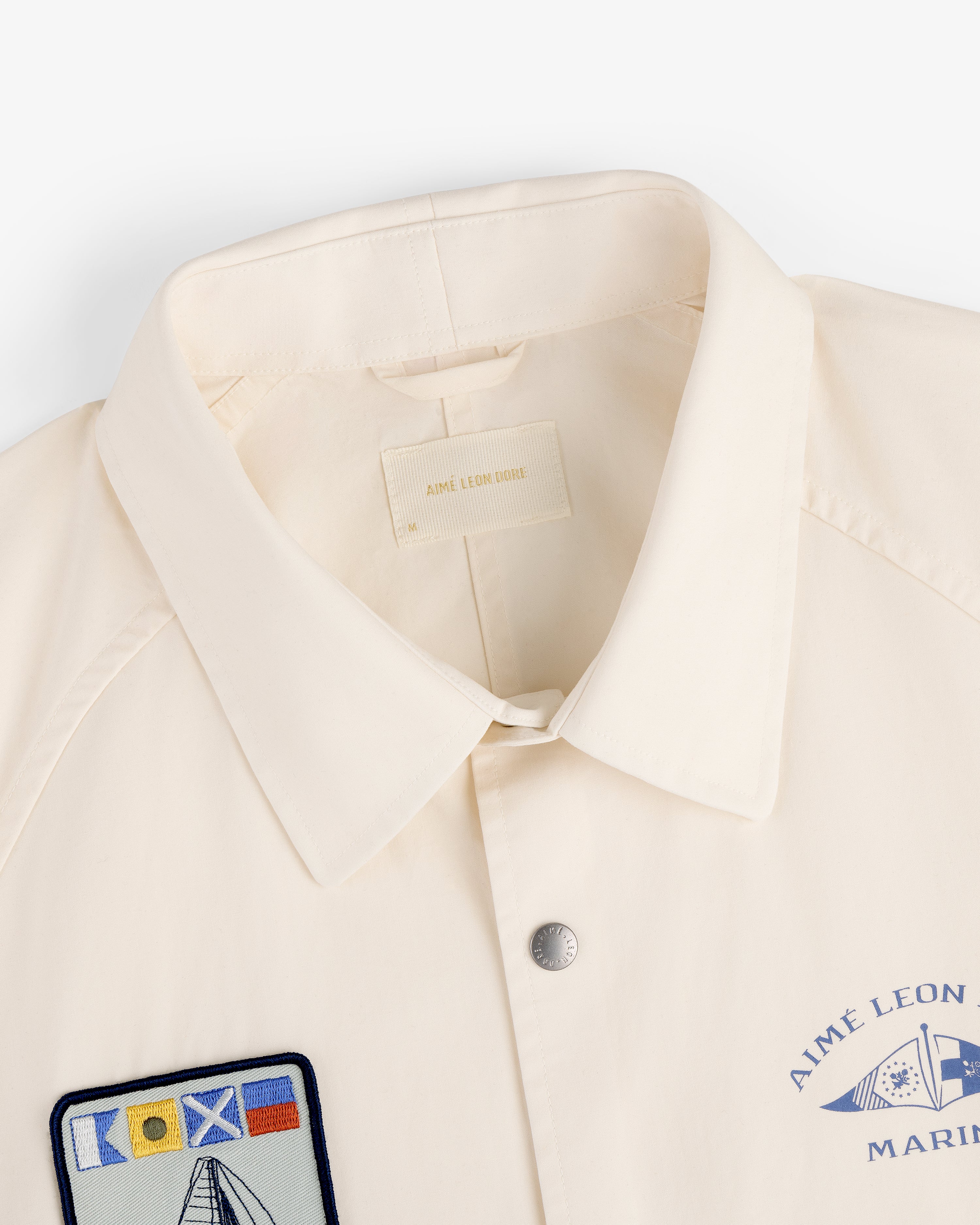 The Marina Jacket is a cream collared jacket with chest patches, featuring a nautical flag and AIME LEON DORE text.