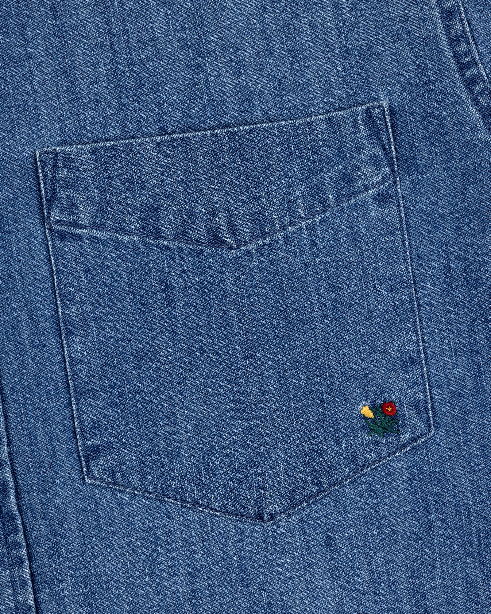 Close-up of the Crest Chambray Shirt's blue denim pocket with a small red and yellow flower embroidery at the corner.