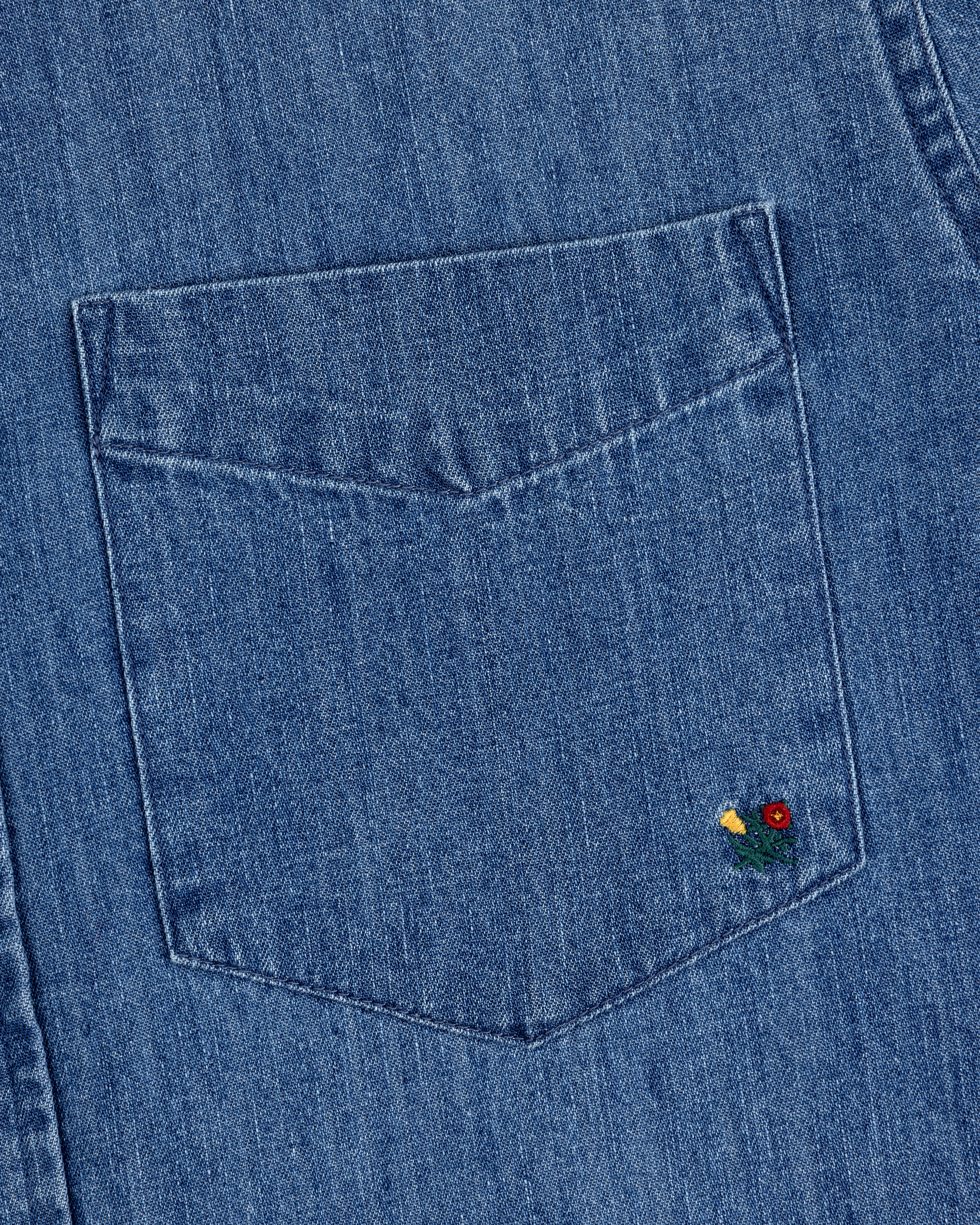 Close-up of the Crest Chambray Shirt's blue denim pocket with a small red and yellow flower embroidery at the corner.
