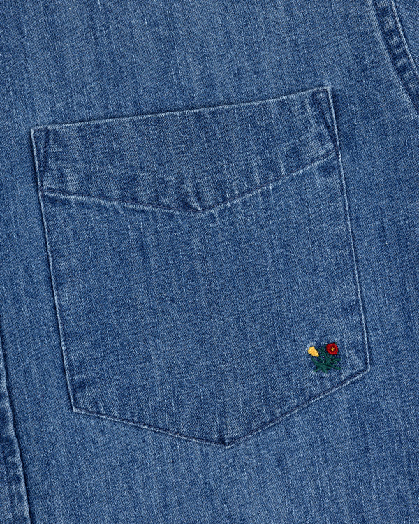 Close-up of the Crest Chambray Shirt's blue denim pocket with a small red and yellow flower embroidery at the corner.
